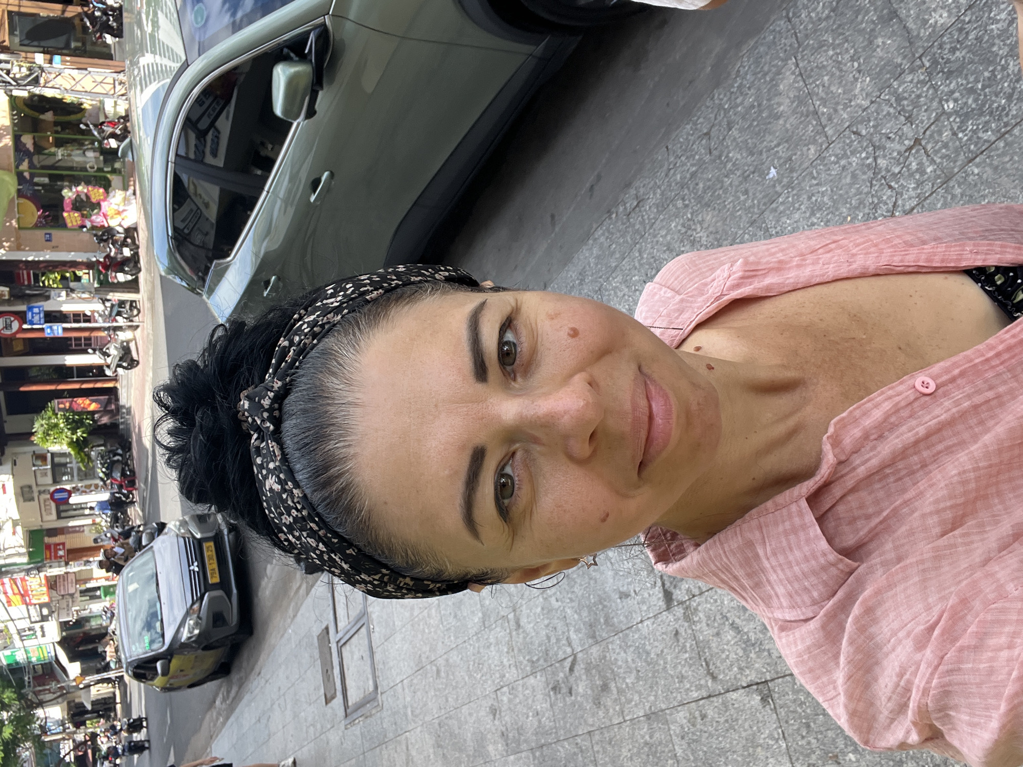 A woman with dark hair, pulled back and adorned with a patterned headband, is taking a selfie on a city street. She is wearing a pink, textured button-up shirt and a black and green patterned top underneath. Her expression is a slight smile. In the background, a gray Mitsubishi Mirage taxi with Vietnamese license plate "79A-130.25" is visible, along with other vehicles and pedestrians. The street is paved with gray tiles, and buildings with awnings line the sidewalk. The scene suggests a sunny day in a bustling urban environment. The presence of the Mitsubishi Mirage taxi with a local license plate, along with the architectural style of the buildings and the general street activity, are characteristic of Nha Trang, Vietnam.
