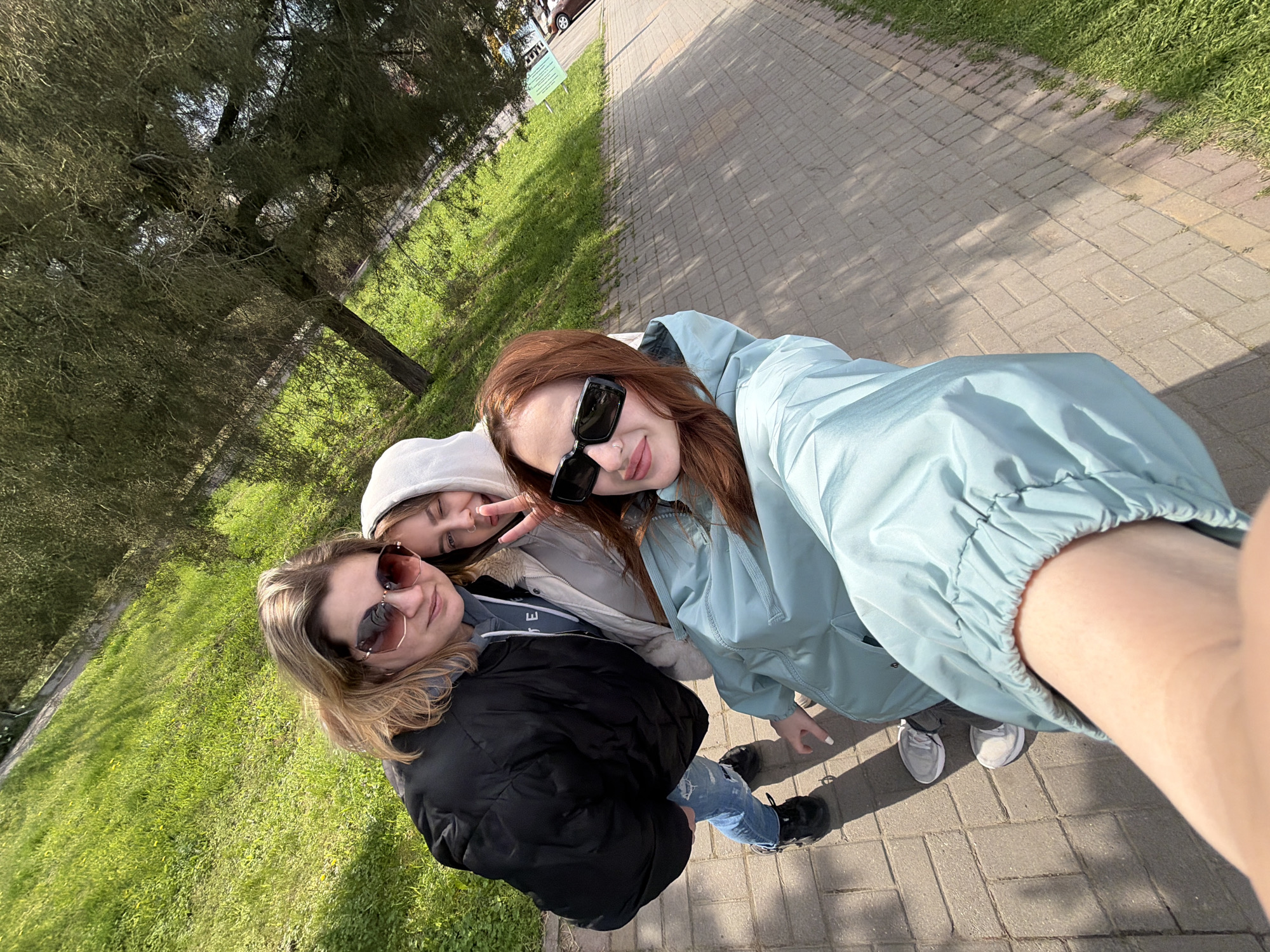 A selfie, taken at an upward angle, depicts three young women standing on a paved pathway. To the left, a woman with blonde hair wears a black jacket and sunglasses. To the top left, a second woman with a white hat makes a peace sign. To the right, a third woman with red hair wears a light blue jacket and sunglasses. All three women are smiling. The pathway is made of grey brick. To the left of the pathway is green grass and a tree. The location is Unknown, Unknown.