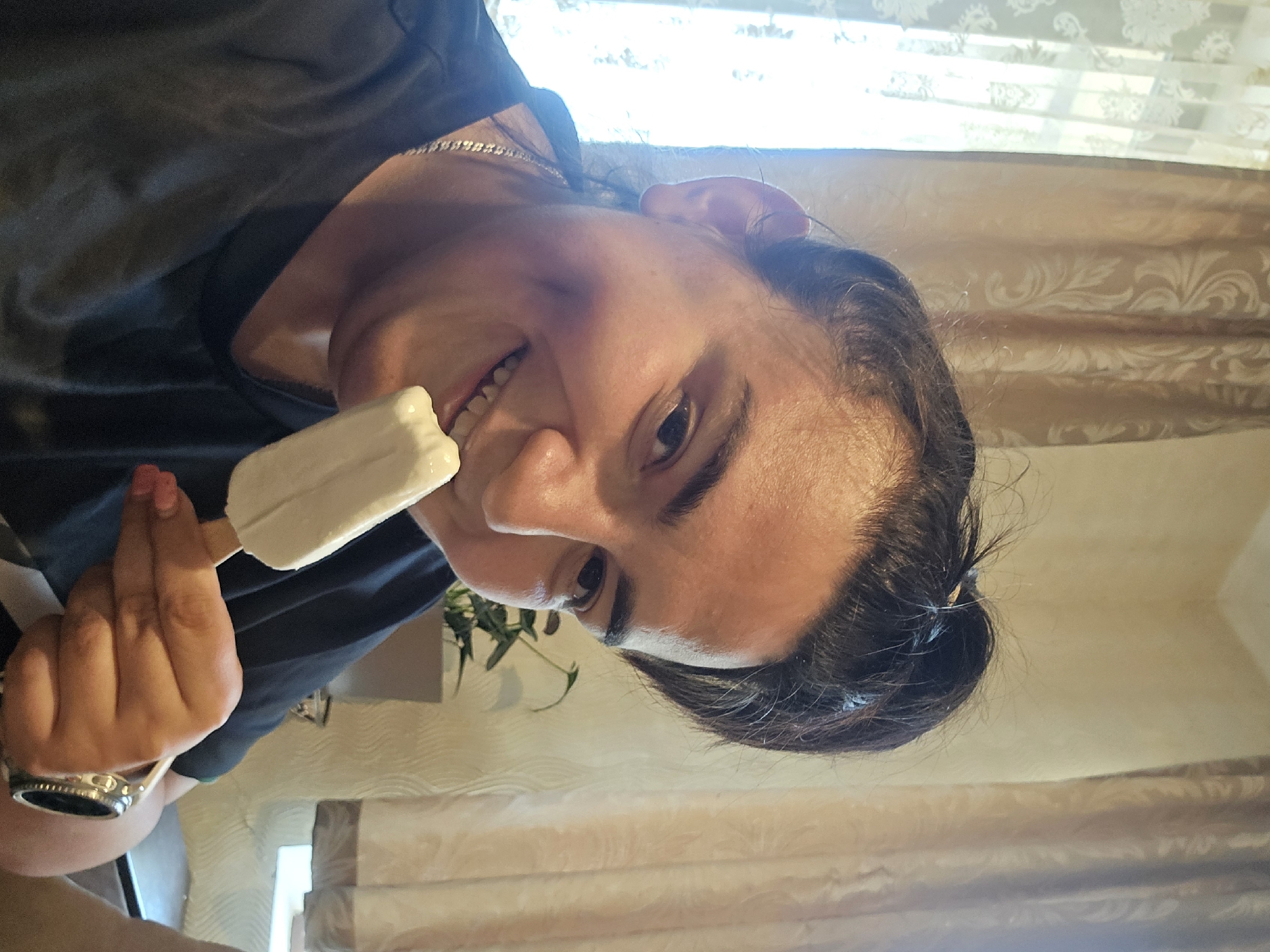 This image captures an individual indoors in Rudnik, Bulgaria, engaged in consuming a white popsicle or ice cream bar. The subject, a person with dark hair styled in a bun, features a visible dimple on the left cheek and is smiling broadly, revealing teeth. They are wearing a dark, short-sleeved t-shirt, a silver-toned chain necklace, and a metallic wristwatch on their right wrist. Pink nail polish is visible on the fingers of the right hand, which holds the wooden stick of the ice cream. The background on the left reveals a window covered by a sheer white lace curtain with a floral pattern, allowing bright natural light to enter. To the right, a drawn light brown or beige curtain with a distinct pattern is visible. A potted plant with dark green leaves is situated behind the individual's right shoulder. The wall behind the subject appears to be a light, neutral color.