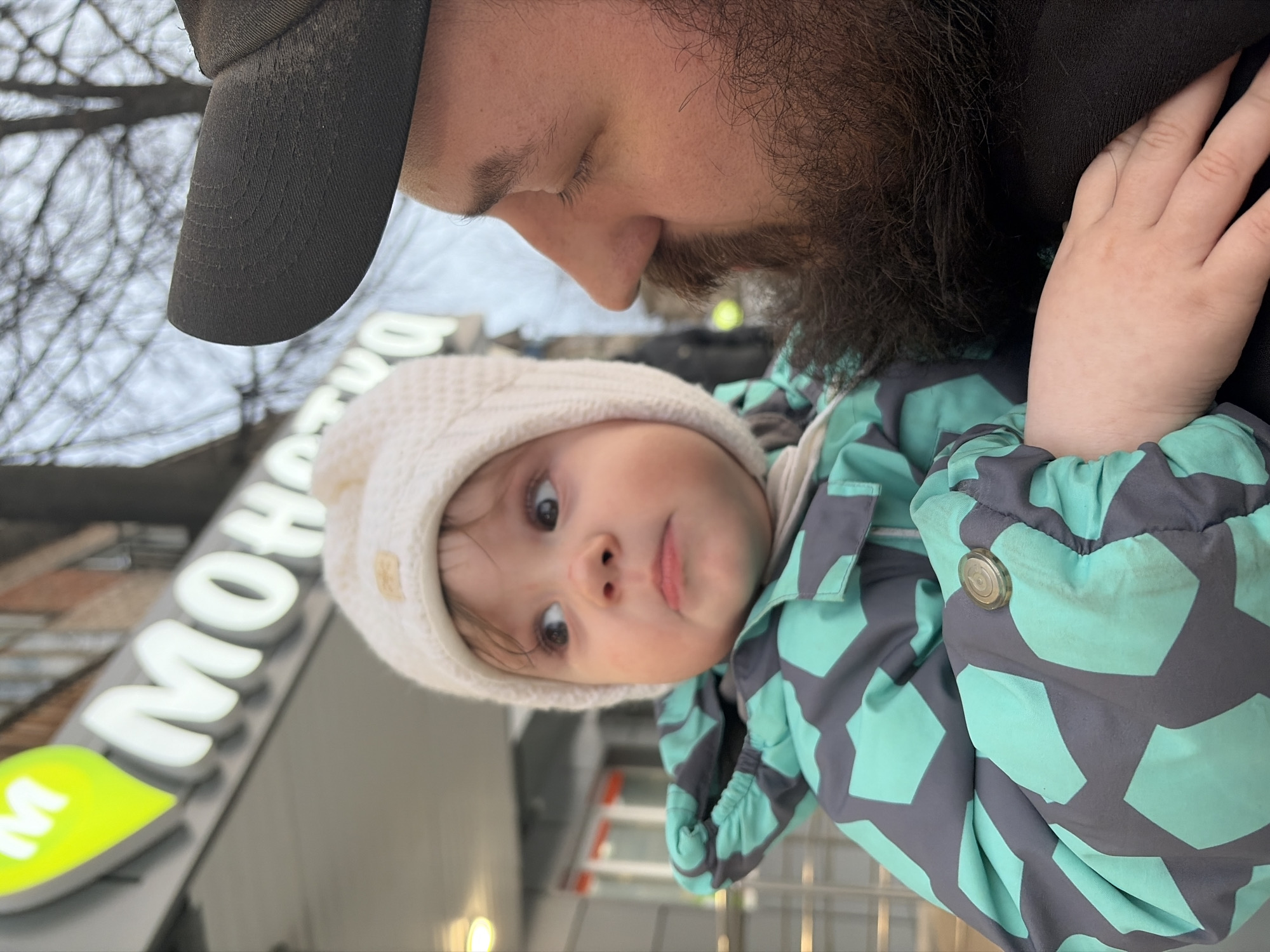 A close-up shot shows a bearded man wearing a dark cap holding a baby in his arms. The baby, dressed in a light-colored hat and a green and grey patterned jacket, looks up with wide eyes. A building with the illuminated word "MOHETOBA" and a green leaf symbol is visible in the background. This building is in Chaikovsky, Russia.
