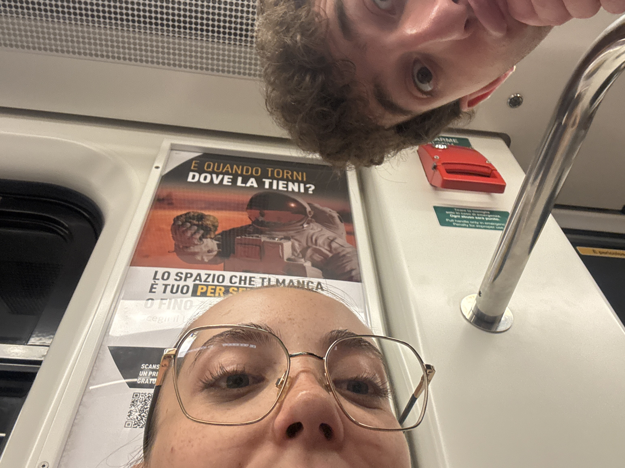 The image appears to be a selfie taken from a low angle inside a subway car. Two people are visible: a person with glasses is in the foreground, looking directly at the camera. Above, another person with curly hair is leaning into the frame, also looking at the camera.
In the background, there is a poster with an astronaut on it, next to an emergency handle. The text on the poster includes "E QUANDO TORNI DOVE LA TIENI?" and "LO SPAZIO CHE TI MANCA È TUO PER". The emergency handle has a sign that says "ARME" and "Tirare la maniglia solo in caso di emergenza. Ogni abuso sara punito. Pull handle only in emergency. Penalty for improper use". There is also a QR code at the bottom of the poster.