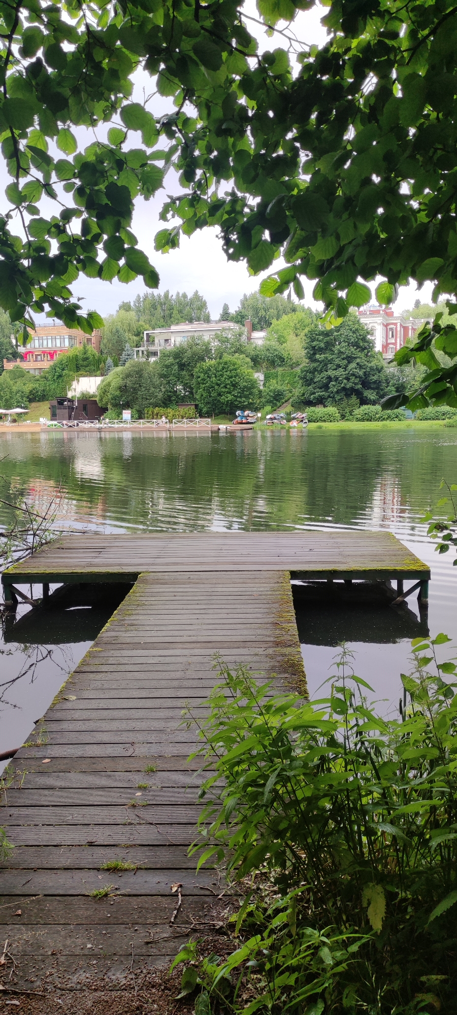 The image shows a wooden pier extending into a calm body of water. Lush green foliage from overhanging trees frames the top of the scene. Across the water, a residential area is visible, featuring several houses with distinct architectural styles, including red-brick and white facades, nestled among dense greenery. Some of the houses appear to be multi-story villas. A small beach area with a few loungers and a pier structure is also present on the far bank, along with several small boats or kayaks docked. The water reflects the overcast sky and the surrounding landscape. The pier itself is weathered and shows signs of age, with some moss growing on its surface. The foreground is partially obscured by tall green plants. The overall scene depicts a tranquil lakeside setting in a suburban or semi-rural environment, likely Angelovo, Russia, given the architectural style and landscape.