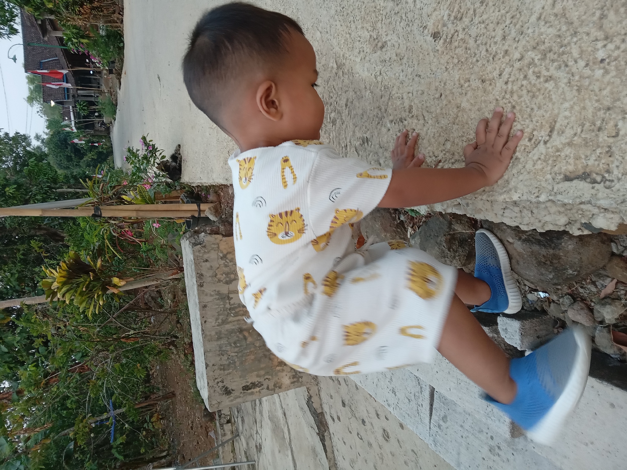 A young child, wearing a white shirt with a tiger pattern and blue sneakers, is on their hands and knees on a textured, light-colored stone surface. The child is leaning forward, with their head down. To the left of the child is a concrete structure and a rocky area. In the background, a paved road leads through a village with traditional Indonesian houses adorned with Indonesian flags. Lush green trees and foliage are visible on both sides of the road and in the background, suggesting a rural or semi-rural setting in Kedunggudel, Indonesia. The lighting indicates it is either early morning or late afternoon.