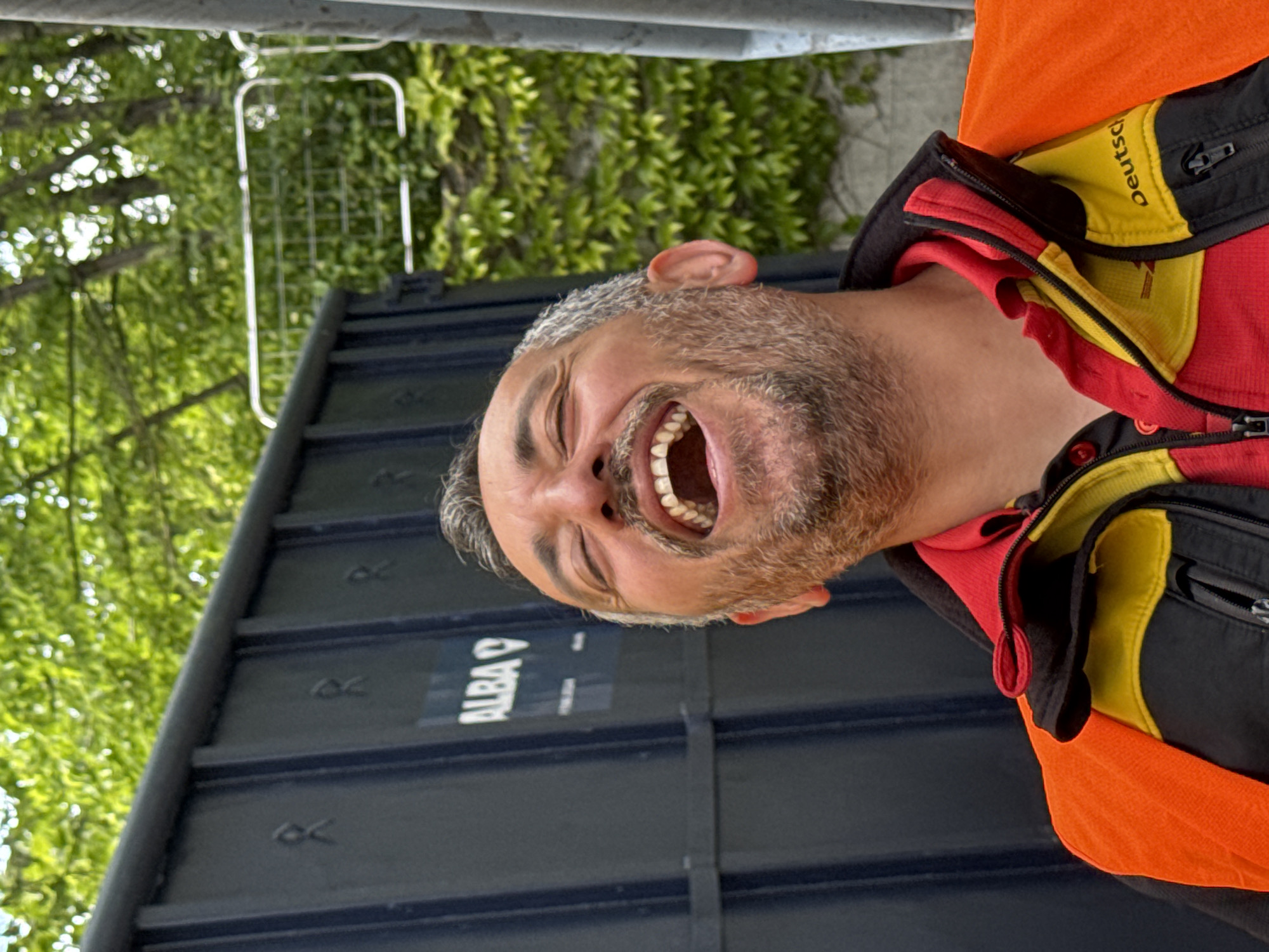 A middle-aged man with graying hair and a beard is laughing, exposing his teeth. He is wearing an orange vest over a jacket with the German flag colors (black, red, and yellow) visible on the collar. The background includes a dark blue metallic structure with "ALBA" printed on it, possibly a container or bin. Behind that, green foliage is visible, suggesting a natural or park setting. The location is Bietigheim-Bissingen, Germany.
