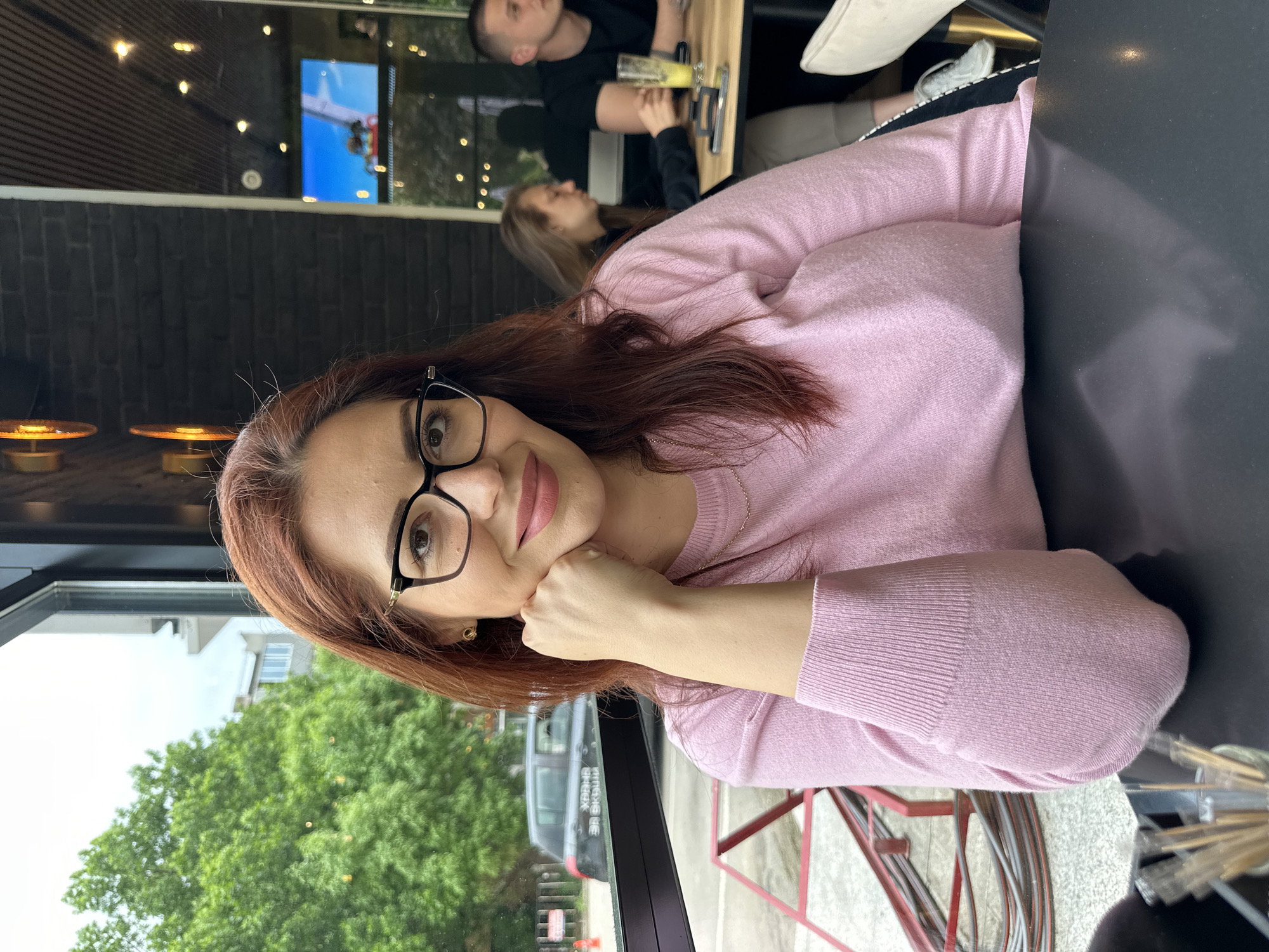 A woman with auburn hair and glasses sits at a table in what appears to be an outdoor or semi-outdoor restaurant or café setting. She is wearing a light pink sweater and has her chin resting on her hand, as if posing for the photo. In the background, through the large window or open doorway, green trees are visible, as is a parked car. A man and a woman are visible behind her, sitting at a table, with a drink in front of the man. The location details, city of Montana, Bulgaria are not verifiable in this image.