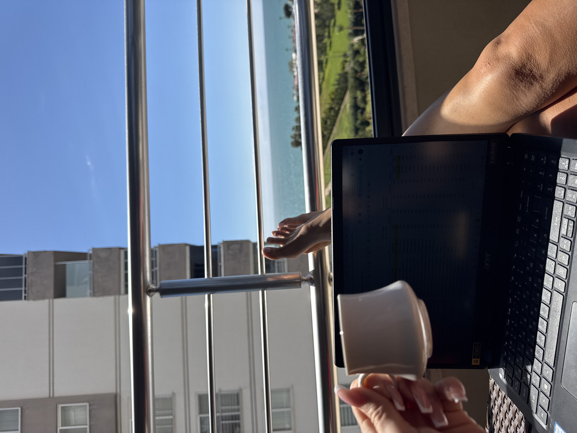 A person is seated on a balcony with a laptop and a mug of coffee, overlooking a beach and an urban landscape, presumably in Erdemli, Turkey. The view through the metal railing shows a coastline with light-colored sand, turquoise water, and a green space adjacent to the shore. The laptop displays a spreadsheet or grid-like document, and the person's leg is visible on the right side of the frame. The foreground includes the person's hand holding the mug and a glimpse of apartment buildings across from the balcony.