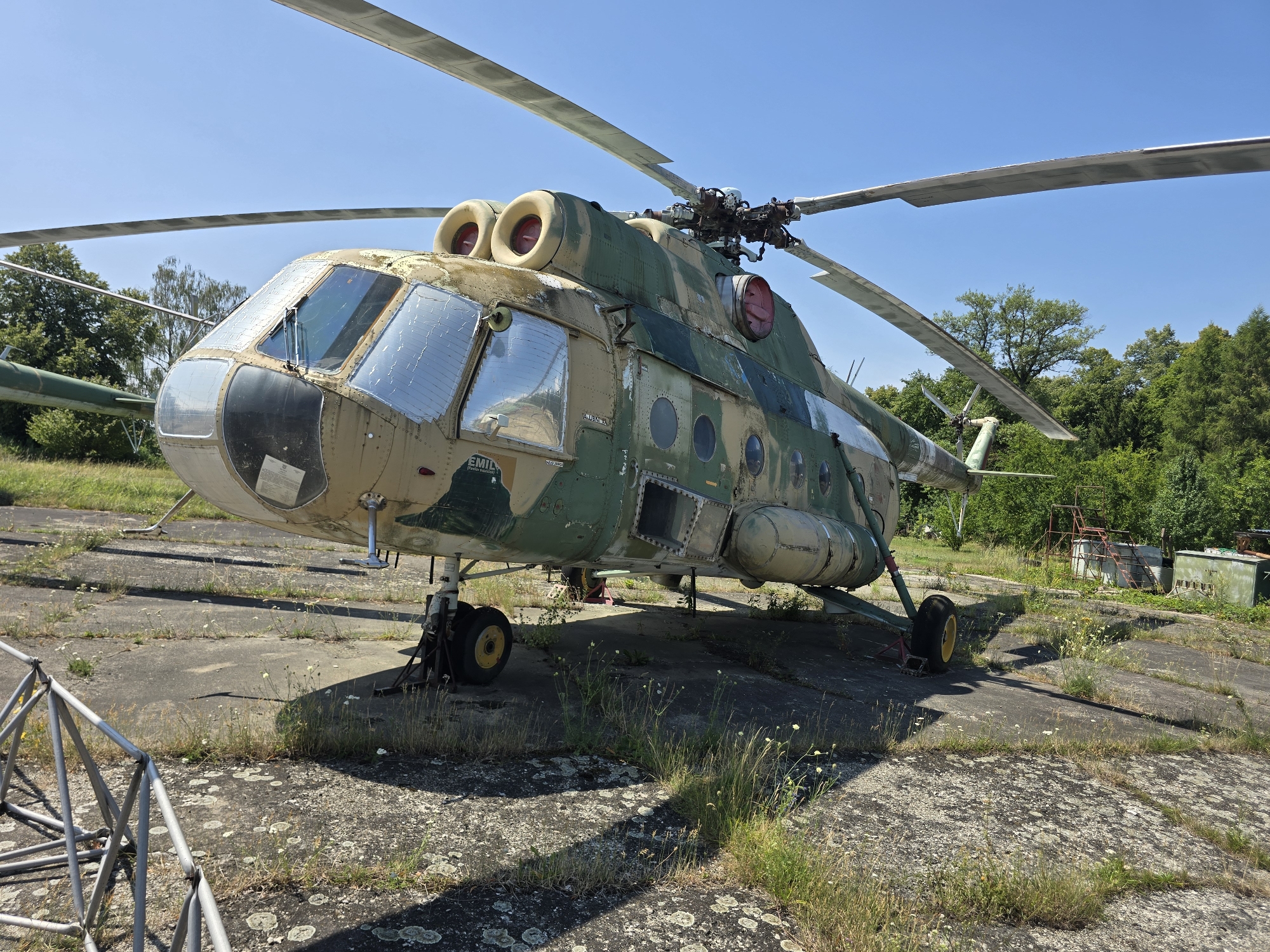 In Pustiměř, Czechia, a grounded military helicopter, camouflaged in green and tan, rests on an outdoor concrete pad. The helicopter, appearing weathered, features visible rotor blades, a cockpit with reflective windows, and various circular porthole-like windows along its fuselage. It is supported by its landing gear, with one wheel clearly visible in the foreground. The surrounding environment consists of overgrown grass, sparse vegetation, and a clear blue sky overhead. In the background, a dense line of trees and several industrial-looking structures are present. The scene suggests a static display or an abandoned aircraft. There are no people visible in the image.