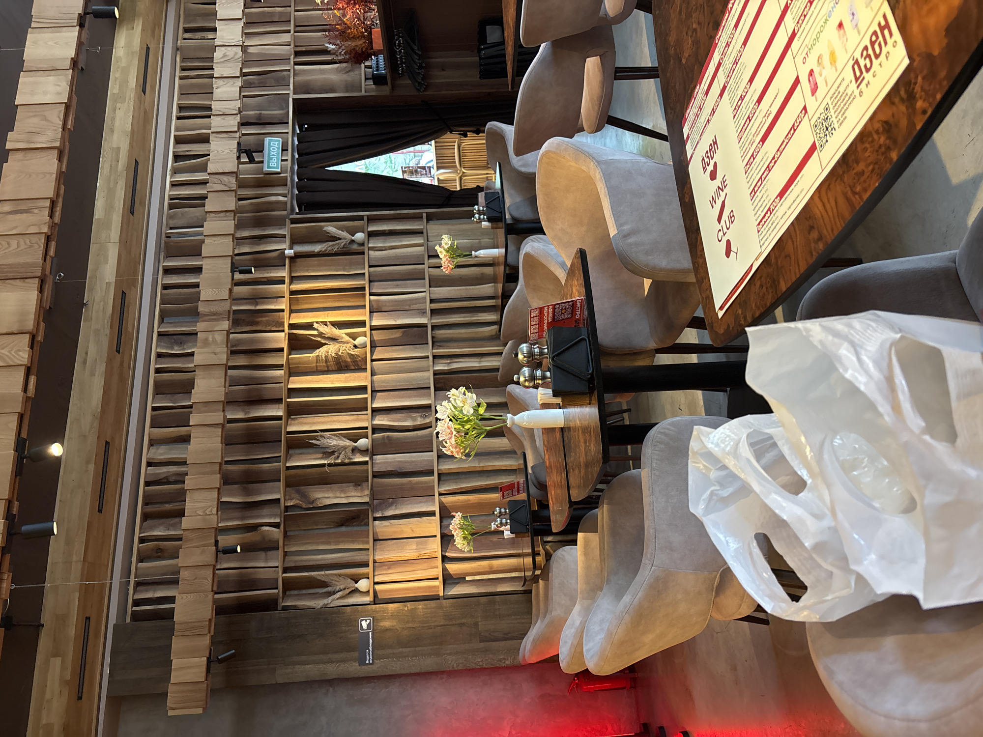 The image depicts the interior of a restaurant or cafe, possibly located in Sochi, Russia. The setting features a dominant theme of natural wood, with walls constructed from vertical wooden planks and horizontal shelves. Several chairs with light-colored upholstery are arranged around dark wooden tables.
In the foreground, a white plastic bag is draped over a chair. A menu is prominently displayed on one of the tables, featuring text in Cyrillic script. Visible text includes "выход" (exit), "WINE CLUB," and menu items such as "Бургер," "Шаурма 550p," "Паста 6500," "Том или рис по-балийски 6500," and "Горячее 700р." There's also text related to "дзен" and "отмороженс" (likely referring to ice cream). Small bouquets of flowers in white vases add decorative touches to the tables.
The lighting suggests it could be daytime, with natural light possibly filtering in from a window, though the overall ambiance seems warm and inviting. The scene appears to be set up for service, with chairs and tables ready for patrons. There are no people clearly visible in the frame, focusing the attention on the restaurant's decor and offerings. The design elements, particularly the wooden elements, give the space a rustic yet modern feel.