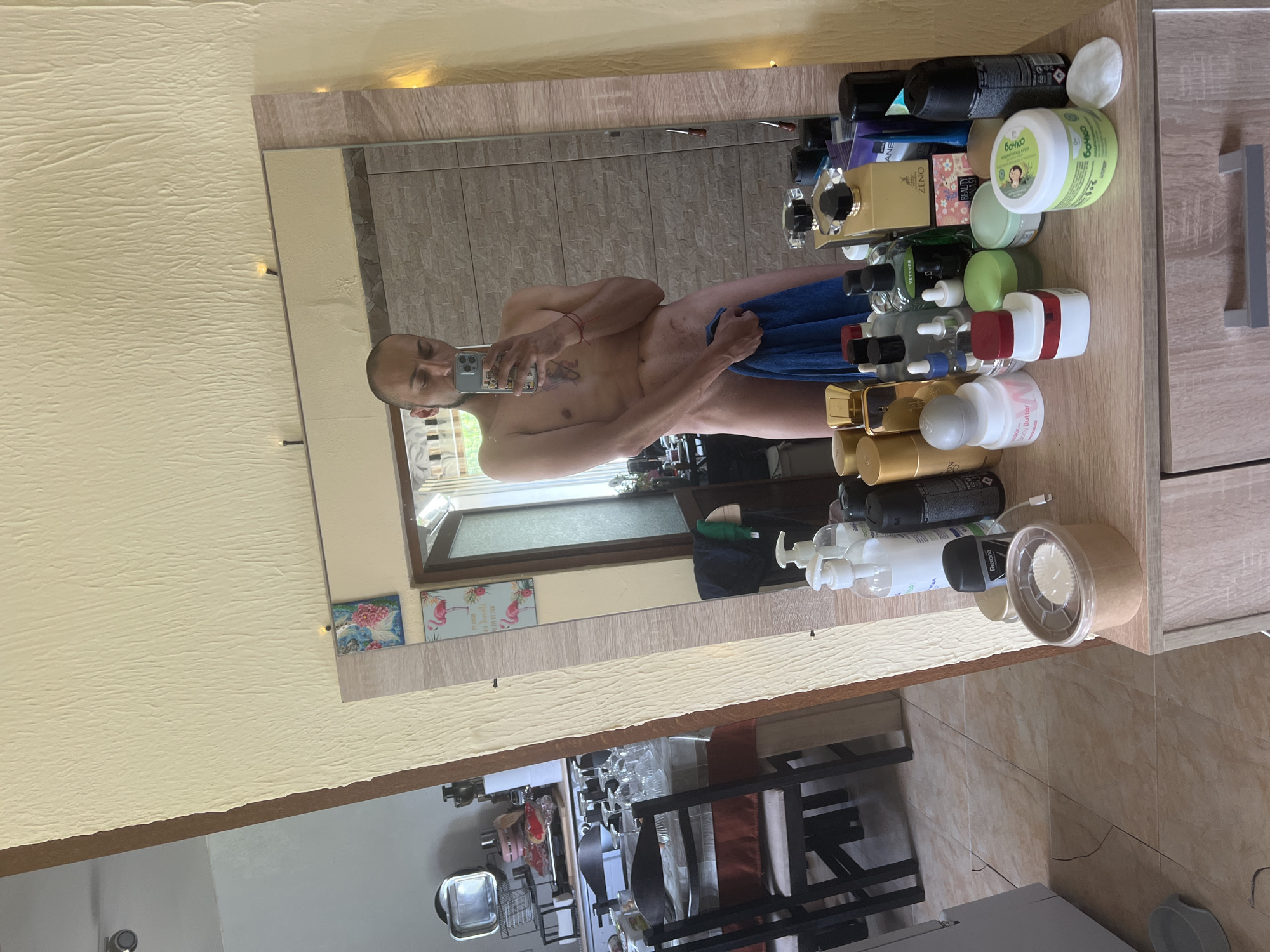 The image shows a naked man holding a blue towel in front of his lower body, taking a selfie in a mirror. He has tattoos on his chest and a red string bracelet on his left wrist. He is standing in a room with a dresser in the foreground, which is covered with various toiletries, including bottles of lotion, spray cans, and jars. In the background, a dining table with chairs is visible, and beyond that, an open doorway leads to another room. The walls are a light yellow color with a textured surface. The scene appears to be an interior of a home, possibly a bathroom or bedroom, given the presence of toiletries and a mirror. The location is not specified in the image or its metadata.
