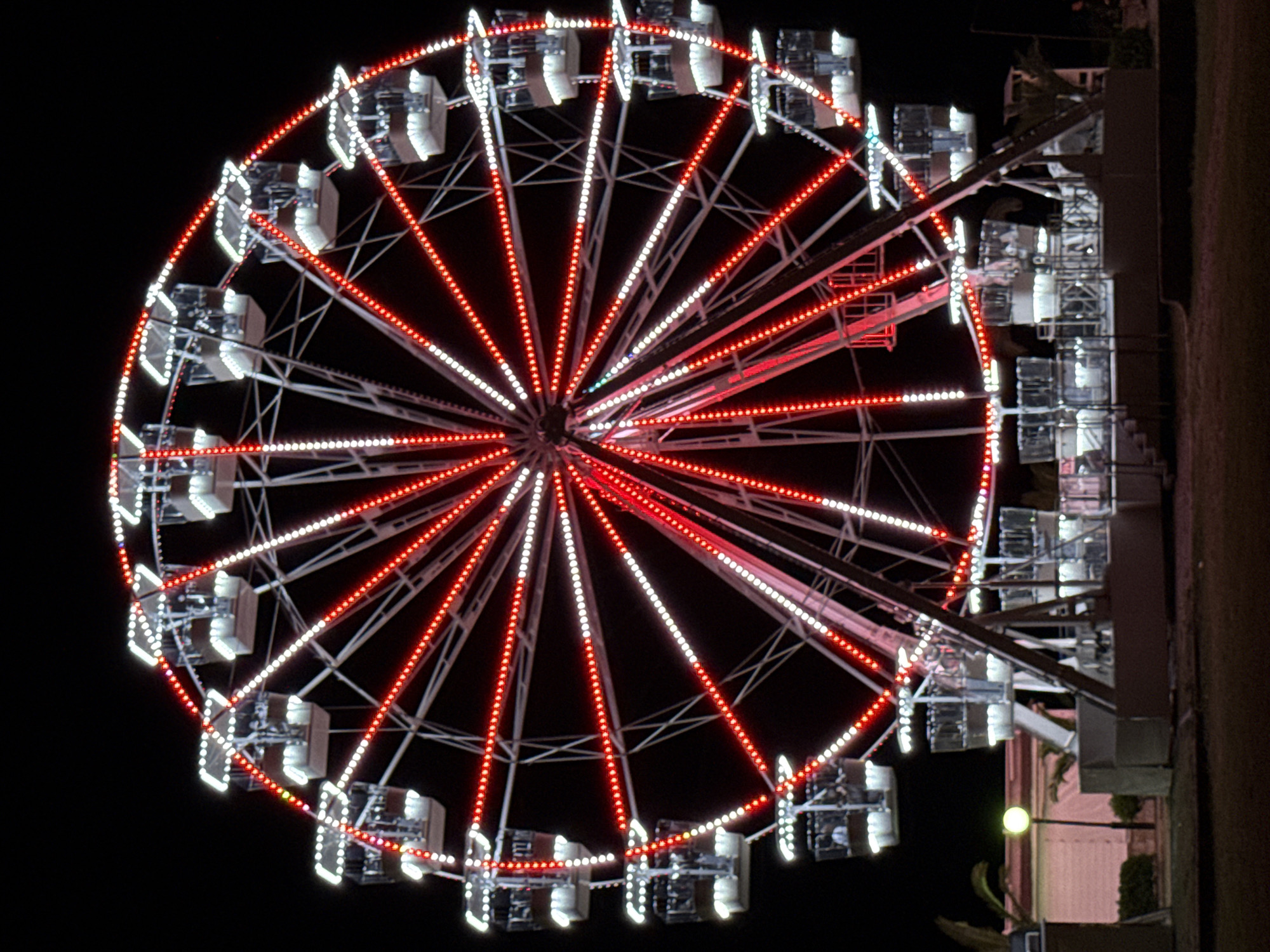 The image shows a Ferris wheel at night. The Ferris wheel is illuminated with red and white lights. It has several gondolas, each with multiple windows. Some gondolas appear to have people inside. The Ferris wheel is situated in a dark, outdoor environment, with some low structures and vegetation visible in the background. A streetlamp is also visible, casting a yellow glow. The ground in the foreground appears to be a dark, grassy area. Based on external knowledge, this Ferris wheel is located on the promenade in Budva, Montenegro.