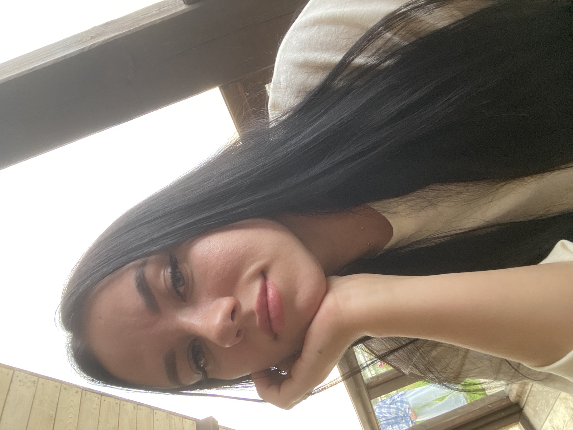 In Chaikovsky, Russia, a young woman with long, dark hair is captured in a selfie. She is resting her chin on her hand, with her arm visible in the foreground. She is wearing a light-colored shirt. The background suggests an outdoor setting, possibly a porch or covered patio, with wooden beams and a bright, overcast sky. In the distance, through a glass pane, a person wearing a blue and white plaid shirt and light-colored pants can be seen standing on grass.
