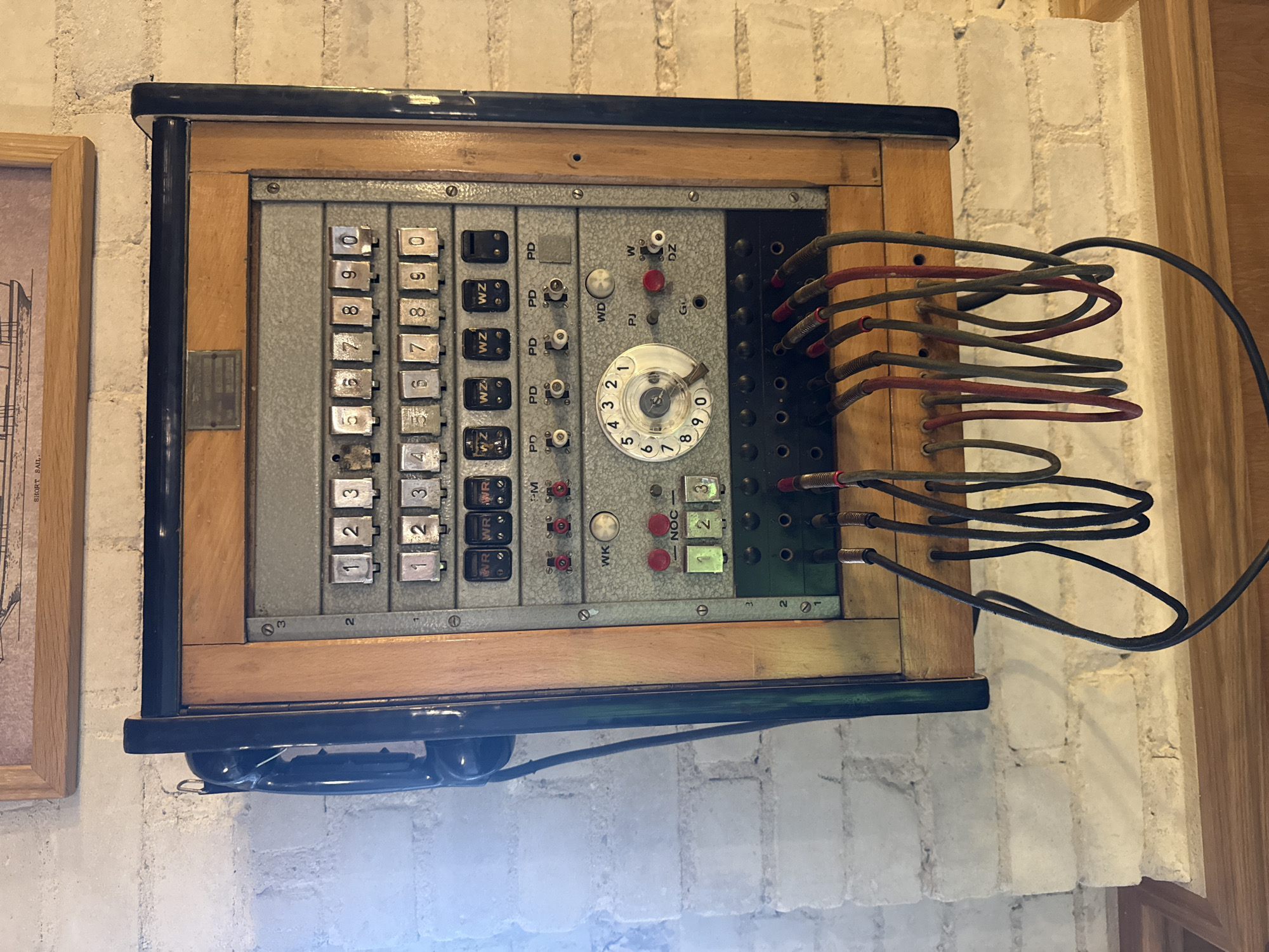 The image shows a vintage switchboard telephone mounted on a white brick wall, possibly in Sława, Poland. The switchboard is made of wood and metal, with multiple rows of buttons, knobs, and a rotary dial. A black telephone handset is attached to the side of the unit. Several cables are connected to the bottom of the switchboard, hanging down towards a wooden surface below. The environment appears to be indoors, with a framed picture visible on the wall above the switchboard. The lighting suggests it is daytime, with natural light illuminating the scene.