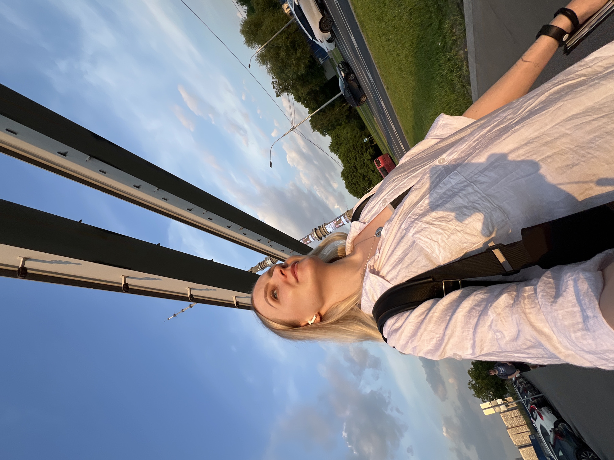 The image is an eye-level shot of a young woman with blonde hair, wearing a white shirt and black bag strap, presumably a backpack. She is looking upwards, potentially at the sky, which is visible in the upper left corner and behind the overhead structure. The location appears to be a bridge or elevated walkway, suggested by the overhead support structure and the cityscape/roadway visible to the right. Cars and greenery are visible on the right, along with additional buildings. The scene may be Khimki, Russia, given the presence of architecture and infrastructure consistent with the urban landscape. The image is shot during the day, likely during the evening.