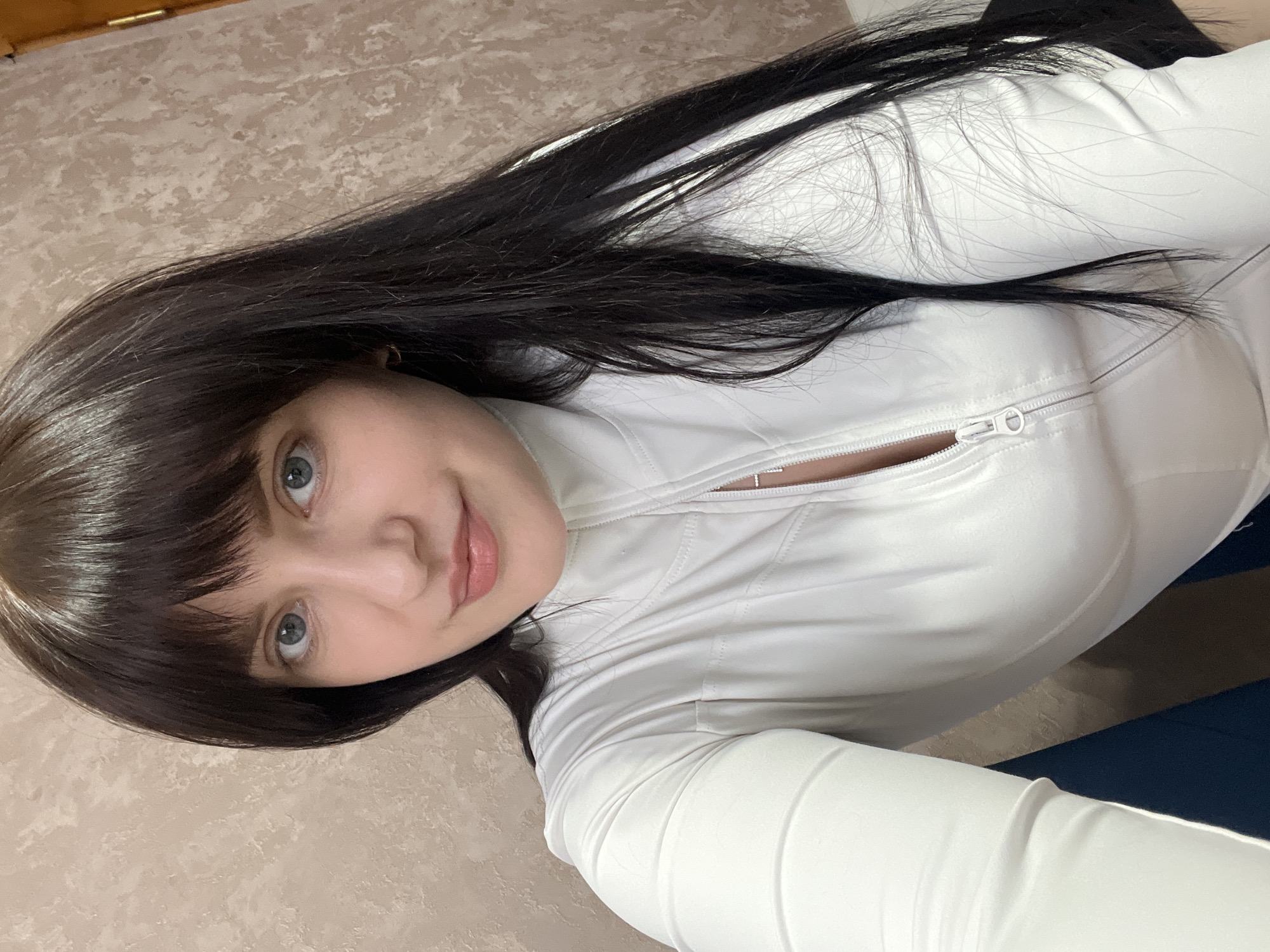 A young woman is captured in a selfie, likely taken indoors in Марковский, Russia. She has long, dark hair styled with bangs that frame her face. Her light-colored eyes, possibly blue or grey, are directed towards the camera, and she has a gentle, soft smile on her lips. She is wearing a form-fitting, long-sleeved white top with a zipper running down the front, which is partially unzipped. The fabric of her top appears smooth and slightly shiny.
The background behind her features a textured, light beige or off-white surface on one side, which could be a wall, bed headboard, or floor. On the other side, a darker, possibly dark blue surface is visible. The overall lighting suggests an indoor setting, and no specific time of day is discernible. The image focuses on her upper body and face, conveying a relaxed and calm mood. There is no visible text in the image.