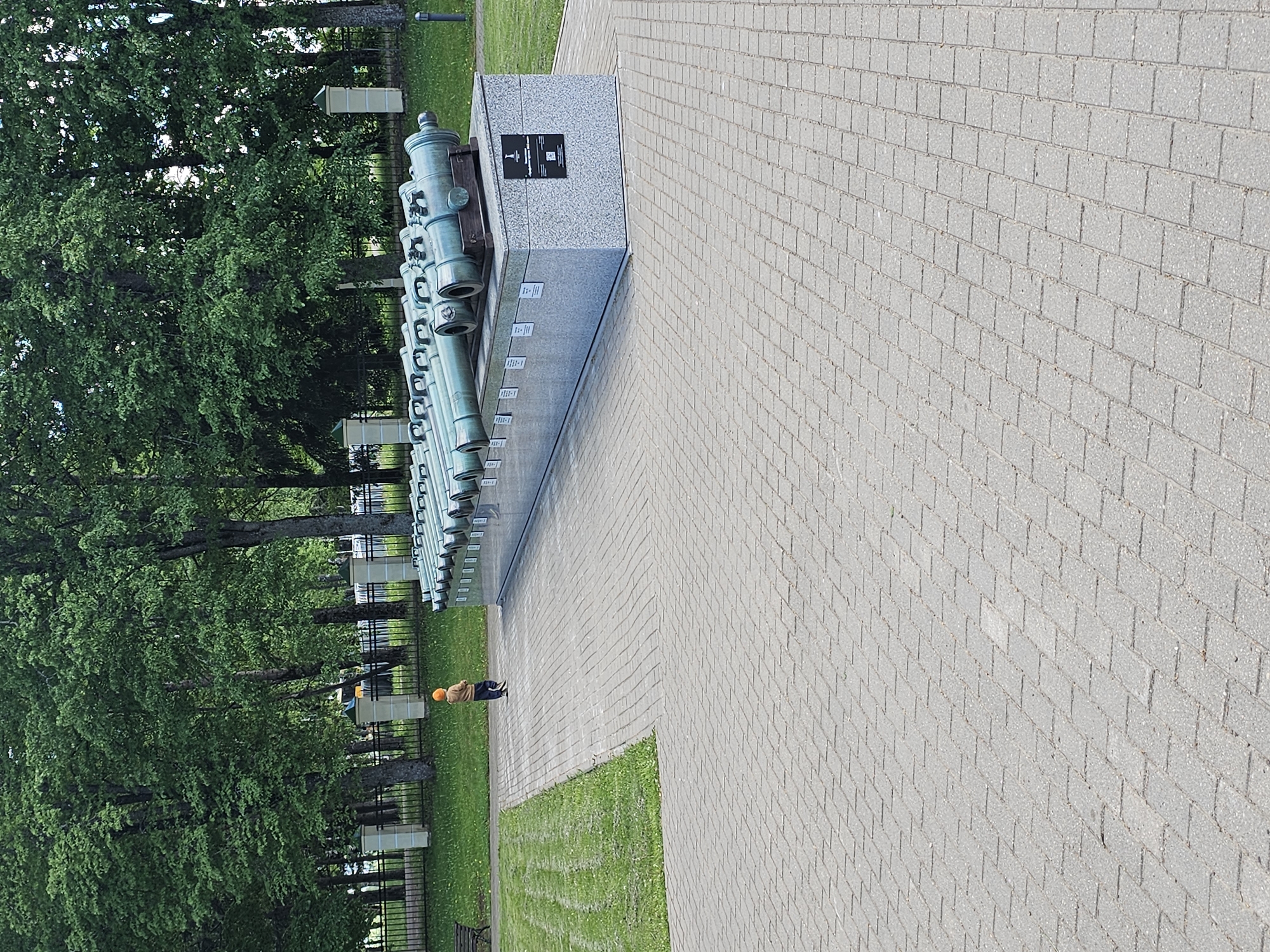 The image shows a row of cannons lined up on a stone base in Borodino, Russia. The cannons are dark green and appear to be made of metal. They are displayed in a line on a raised platform made of gray stone or concrete. A granite slab is affixed to one end of the row with writing on it. A paved brick walkway extends from the cannons. Green trees line the cannons in the background, partially obscured by a fence. A person wearing an orange hat and dark clothing is standing near the cannons. A grassy area separates the cannons from the walkway.