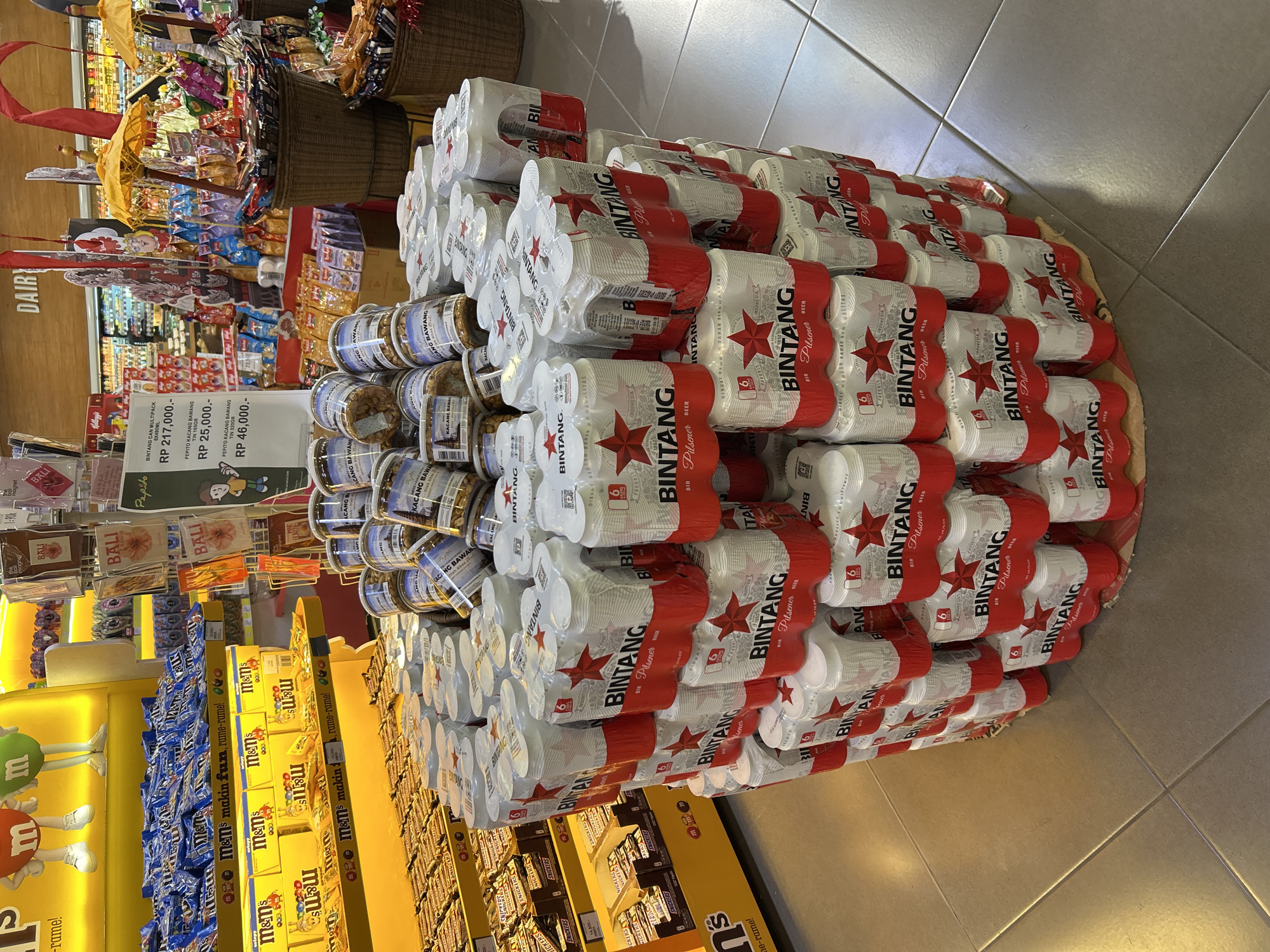 The image shows a retail display of canned Bintang beer in a store, likely a supermarket, in the Ubud District of Indonesia. The display is a circular tower of multipacks of the beer, with each multipack containing six cans. The cans are predominantly white with red branding, featuring the Bintang star logo and text in Indonesian. In the background, shelves are stocked with various grocery items, including M&M's candies and other packaged goods. A price tag is visible on a nearby display. The floor is tiled. There are no people visible in the immediate foreground of the display.