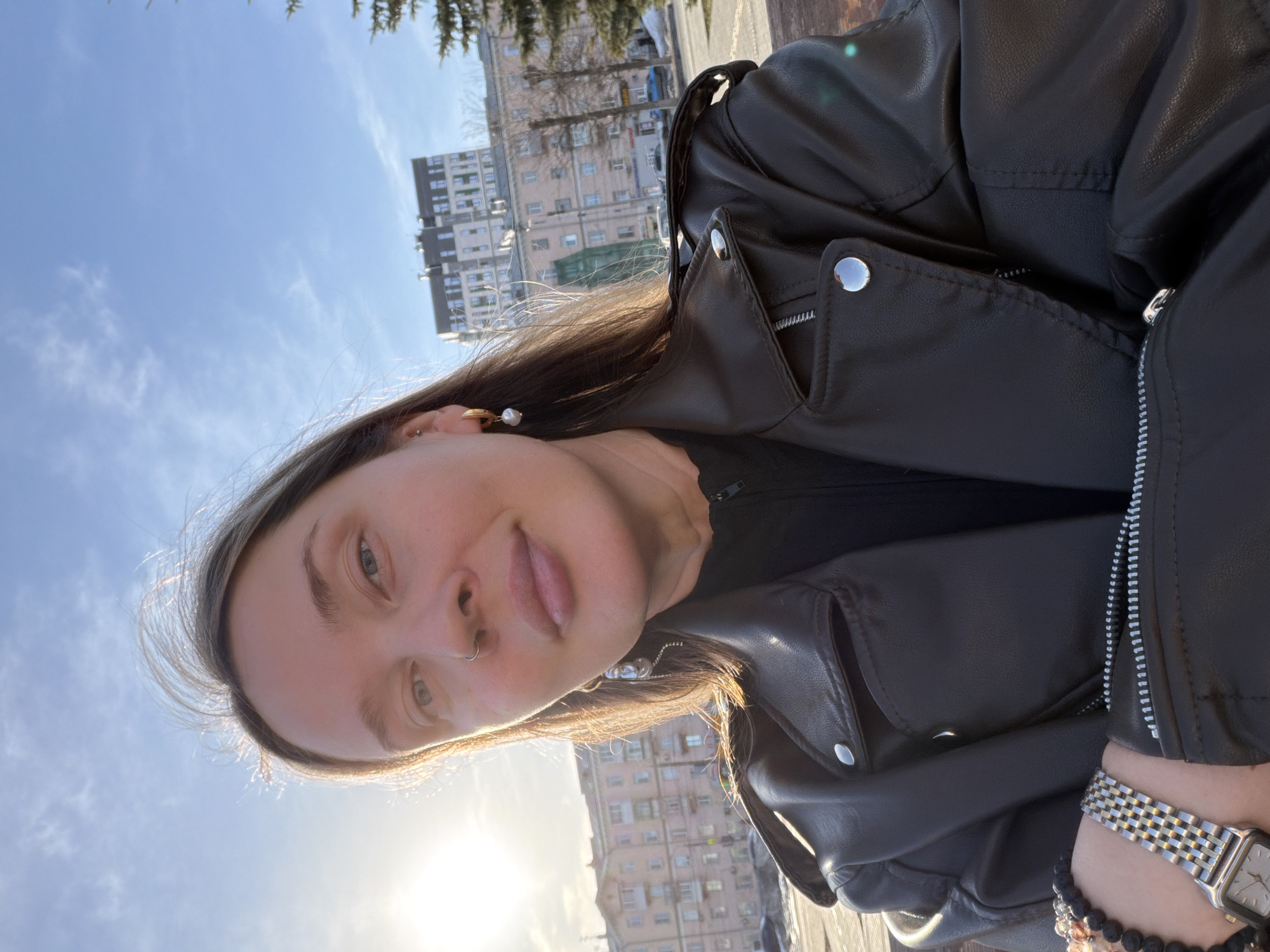 A woman is posing for a selfie in the city of Izhevsk, Russia. The subject has fair skin, light brown hair, and is wearing a black leather jacket, earrings, and a nose ring. She is also wearing a watch and bracelets. Behind her are buildings and trees, with a bright sky visible in the background.