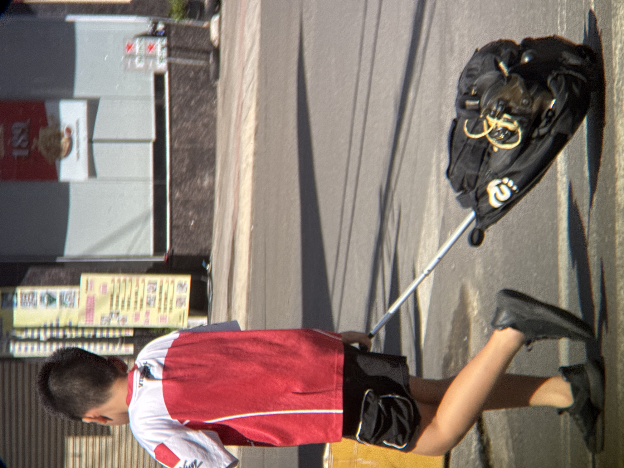 A young boy, seen from the back, walks on a sidewalk in Nha Trang, Vietnam, pulling a black rolling suitcase. He wears a red and white short-sleeved shirt and black shorts. His right leg is forward, and he is looking ahead. The suitcase is large and black with yellow and white markings. In the background, there are buildings with signs, a closed metal shutter, and a grey pavement. The scene appears to be a sunny day.