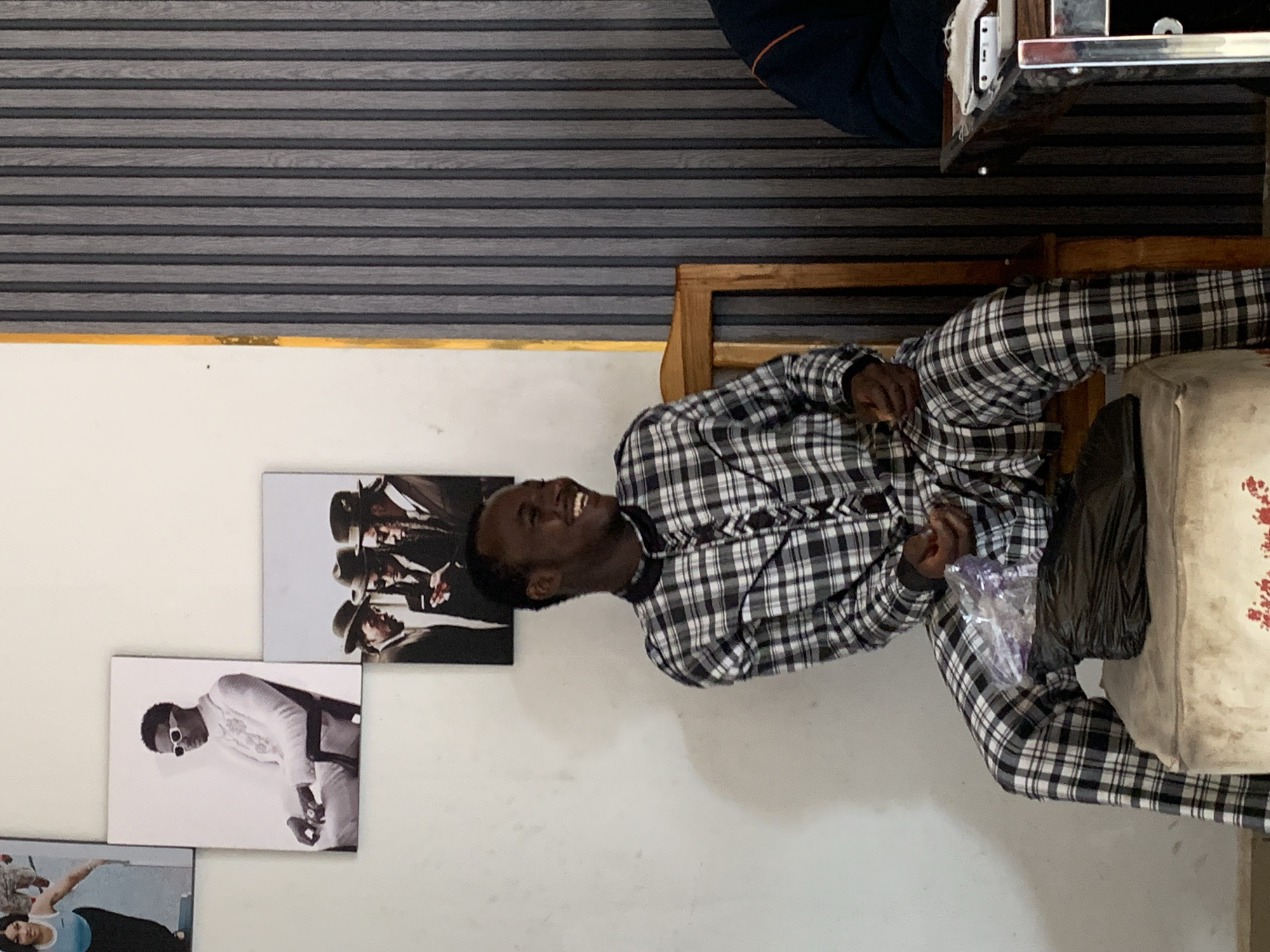 A man is sitting on a chair in an indoor setting. He is wearing a plaid shirt and plaid pants. He is smiling and appears to be interacting with someone out of frame. Next to him on the chair is a plastic bag and a black object. Behind him on the wall are three framed pictures of men. The wall is off-white. Above the wall is a horizontal striped wall covering. Part of a table and another person are visible at the top of the frame. There is no information in the image to suggest the location is Jos, Nigeria.