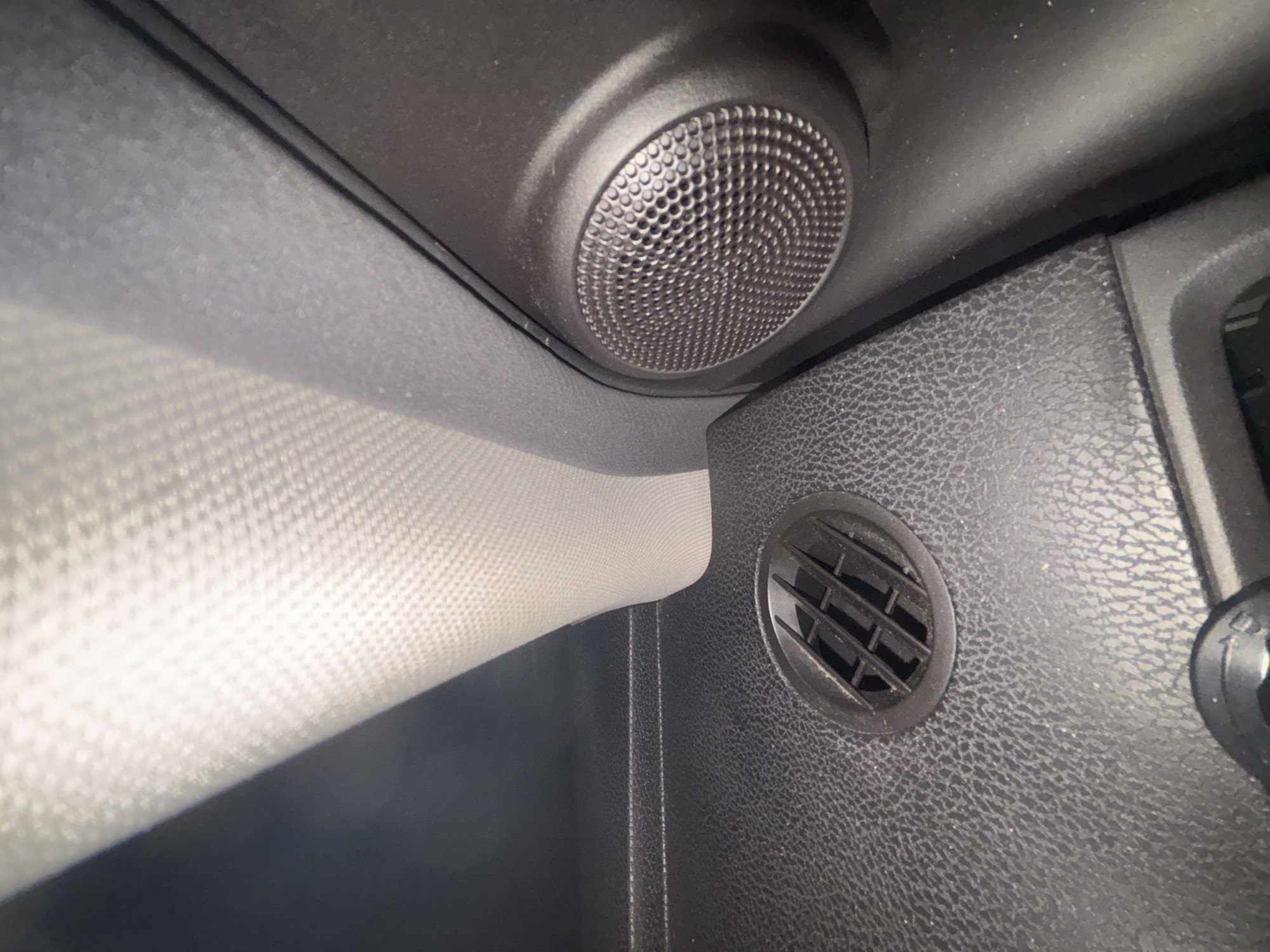 The image shows a portion of a car's interior. Visible are a round speaker grill in the upper portion of the image, a glove compartment, and a small round air vent. The materials appear to be primarily black plastic. The location is not discernible from the image.