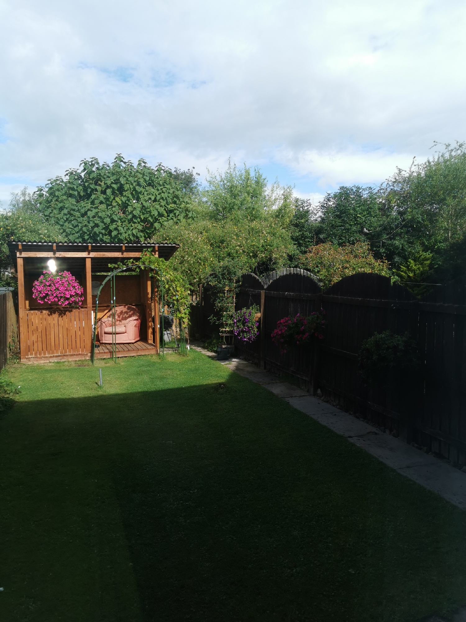 A garden with a wooden gazebo containing a pink armchair and a hanging basket of pink flowers. A dark wooden fence runs along the right side of the image, with more hanging baskets of flowers. A lawn occupies the foreground, with trees and bushes surrounding the garden. The sky is partly cloudy. This scene appears to be in a residential area of Ripon, United Kingdom.