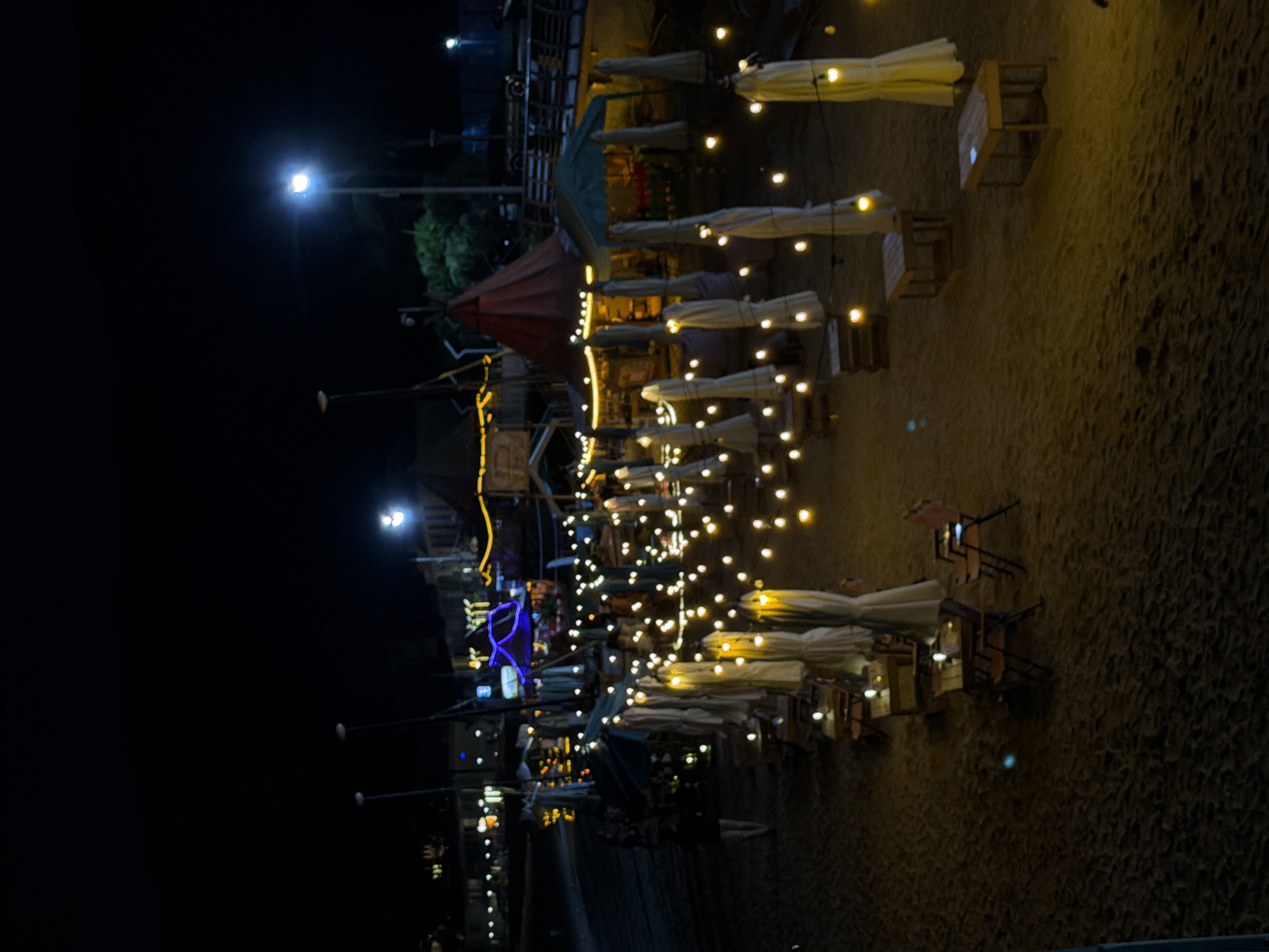 The image depicts a nighttime scene on a sandy beach, likely a commercial area with outdoor seating. Rows of white, closed umbrellas are set up, interspersed with small wooden tables and chairs. String lights with yellow bulbs illuminate the scene, creating a warm and festive atmosphere. In the background, there are structures resembling tents or small buildings with illuminated signage and decorative lights, suggesting a market or food stall area. A few people are visible, including one person walking in the lower left of the frame. The overall impression is that of a vibrant, illuminated beachfront at night, possibly in a tourist or recreational zone within Longgang District, China.