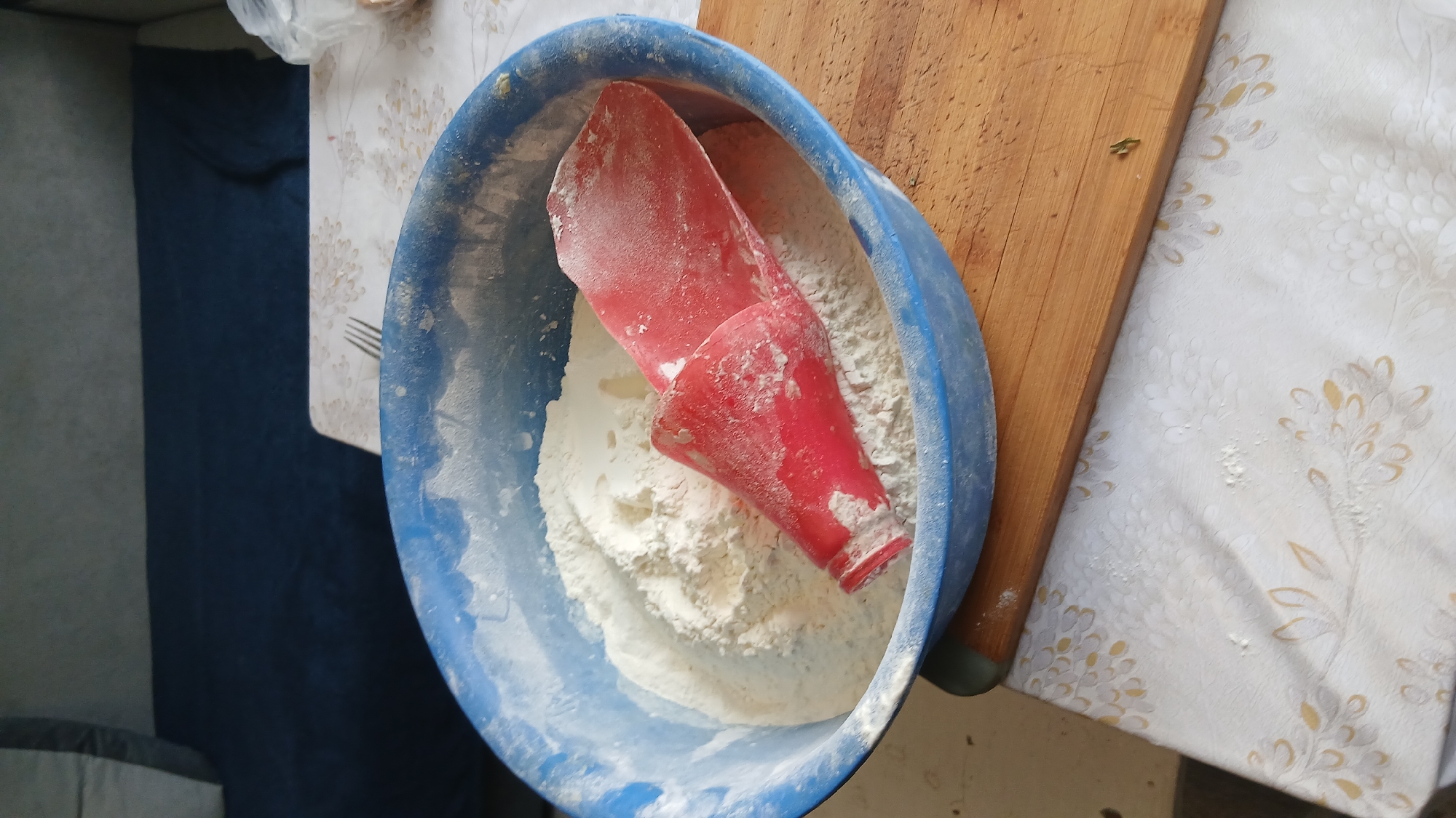 The image shows a blue plastic bowl filled with white flour, with a red plastic measuring cup/scoop embedded inside. The bowl sits on a table next to a wooden cutting board, and the tablecloth has a floral pattern. The scene appears to be indoors. The location, Abzakovo, Russia, is provided as metadata, but is not directly visible in the image.