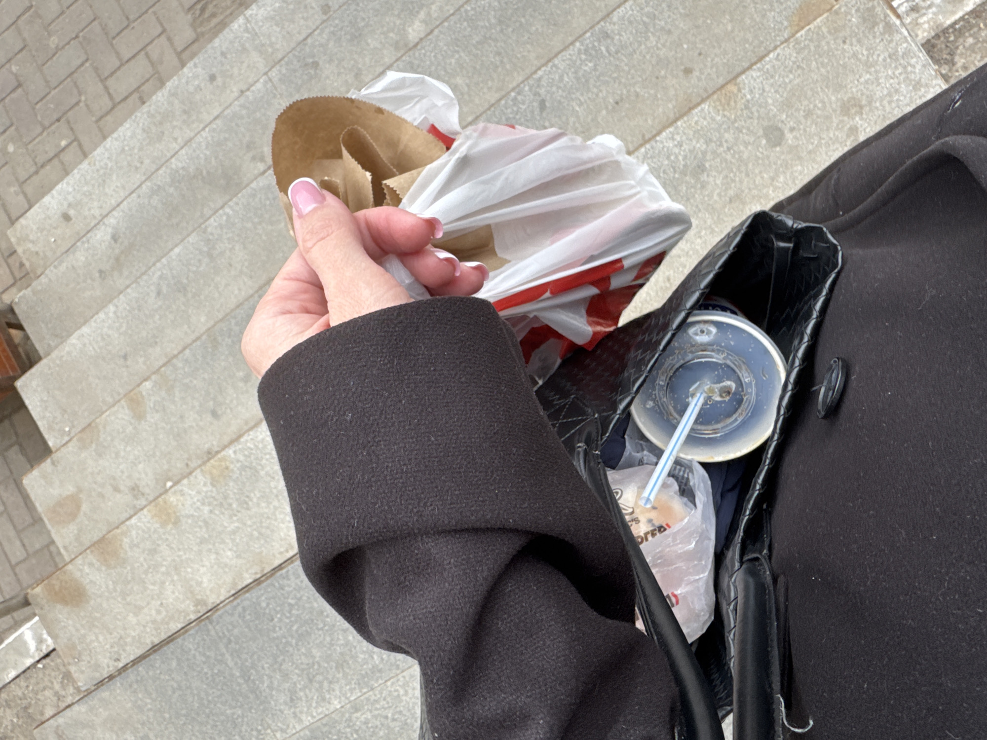 A first-person, downward-angle shot captures a person standing on or near concrete steps in an outdoor setting in Chaikovsky, Russia. The individual's right arm, clad in a dark, possibly black, wool or felt-like coat, extends towards the left. Their hand, with neatly manicured nails featuring a pink and white French tip, gently holds a small, empty brown paper bag.
To the right, a black handbag with a distinctive woven texture is partially open, revealing its contents. Inside, a clear plastic cup with a lid and a blue-and-white striped straw is visible, suggesting a beverage. Tucked beside the cup is a crumpled white plastic bag with red details and partial text "D's" and "OFFEE," hinting at a fast-food or cafe origin, possibly a coffee cup from a chain like McDonald's. Another small, transparent plastic bag is also visible within the main handbag.
The background features light gray concrete or stone steps, showing some minor stains or damp patches. A narrow strip of patterned paving or cobblestone is visible to the far left. The lighting is diffused, suggesting an overcast day. The overall scene depicts a casual moment, possibly after making a purchase or during a break while out and about.