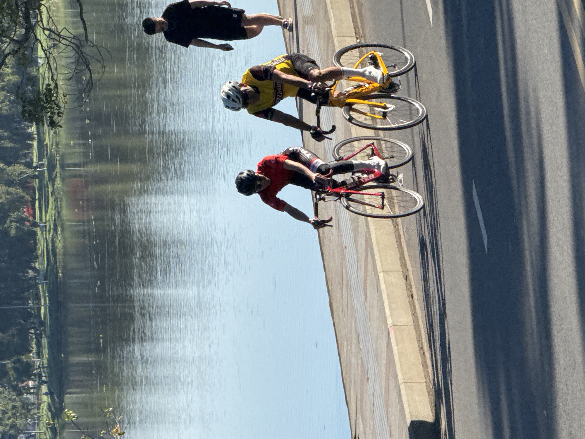 This image captures a scene on a paved pathway next to a body of water, likely Xuan Huong Lake in Da Lat, Vietnam, given the terrain and vegetation. Two individuals are actively engaged in cycling. The cyclist in the foreground is wearing a red jersey and black cycling shorts, with a black helmet. The cyclist to their right is wearing a yellow and black jersey, black shorts, a white helmet, and sunglasses. Both cyclists are on road bikes, with the yellow-jersey cyclist on a yellow bike and the red-jersey cyclist on a red bike. Their bodies are in a bent-over cycling posture, indicating movement. In the background, a third person, dressed in a black t-shirt and black shorts, is walking away from the camera. The environment features a calm body of water reflecting the sky and surrounding trees, a paved walkway with a distinct border, and a road surface in the foreground where shadows of the cyclists are cast. The overall lighting suggests a sunny day.