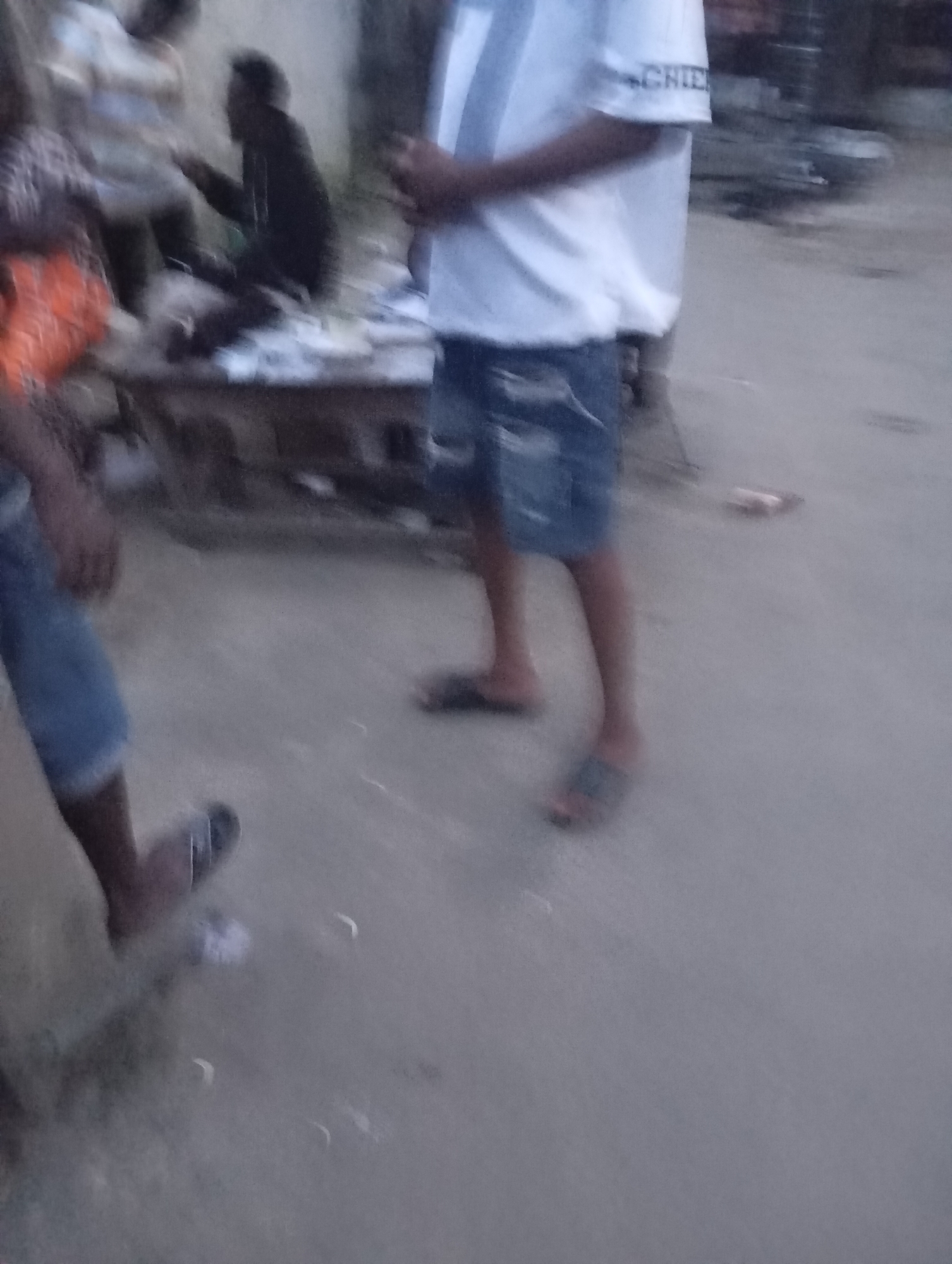 In a dimly lit, outdoor setting, possibly a street or market area in Port-Harcourt, Nigeria, several individuals are present. One person, wearing a white t-shirt with a dark stripe and blue denim shorts, stands in the foreground, facing towards the right of the frame. Another individual, partially visible on the left, appears to be wearing a blue garment and a colorful orange shirt underneath. Further back, a person in a dark jacket is seated. The ground is paved with a sandy or dusty surface. The overall atmosphere is somewhat blurred, suggesting a candid or low-light photograph.