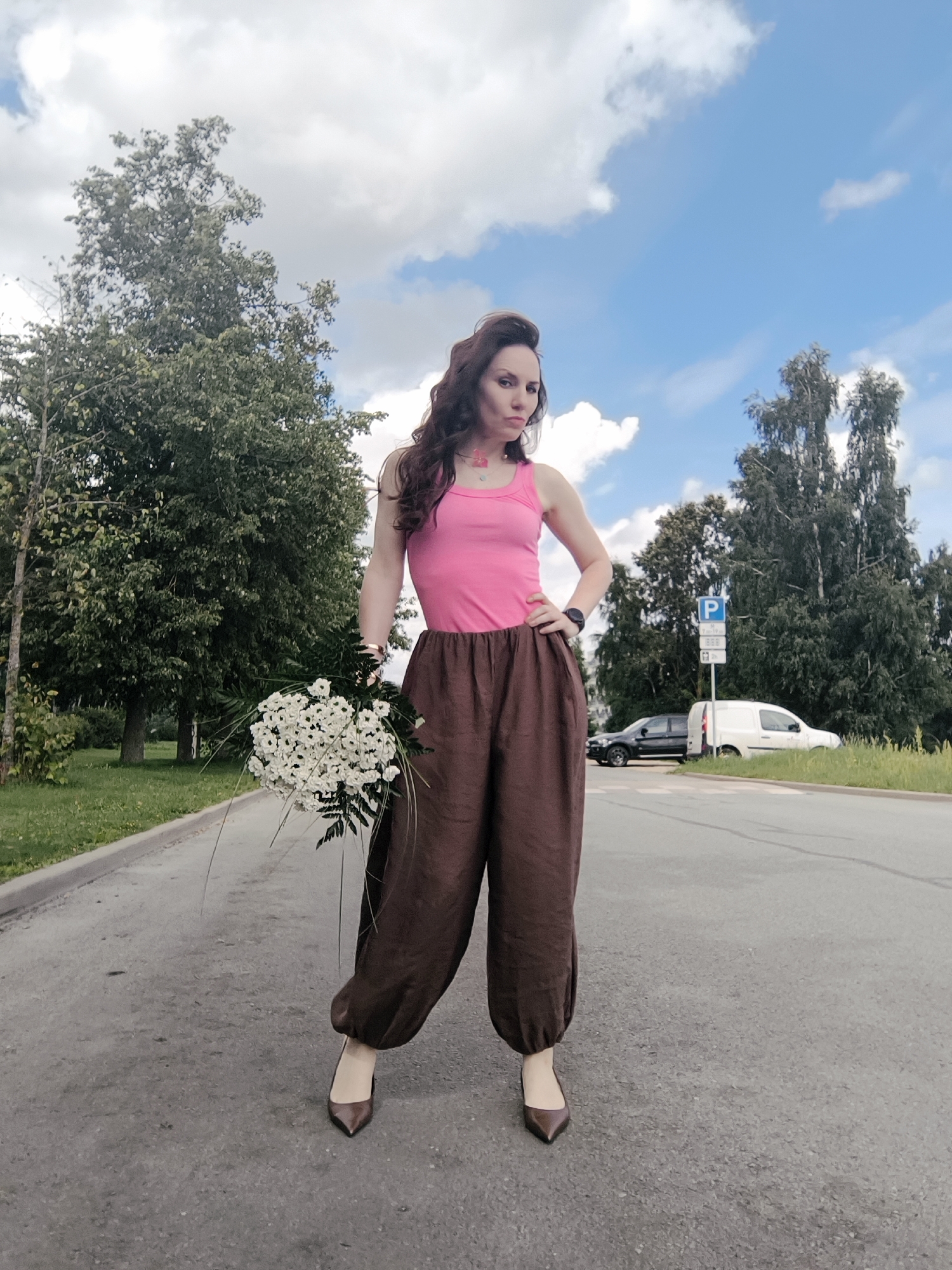 A full-body, eye-level shot depicts a woman standing on a paved road, holding a bouquet of white flowers. She is wearing a pink tank top and loose-fitting brown pants that gather at the ankle. Her brown hair is styled in waves, and she is wearing brown pointed-toe high heels. Her left hand is on her hip, and her right hand is holding the bouquet. In the background, trees and a blue sky with white clouds are visible. To the right of the woman, there is a parking sign and two vehicles parked on the side of the road. The scene appears to be in a suburban or rural area, possibly on the outskirts of Riga, Latvia.
