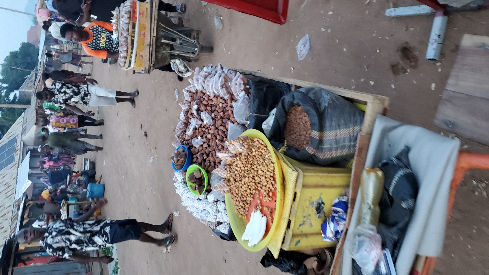 This is a vertical, eye-level, full-shot image capturing a street scene in Jalingo, Nigeria. The foreground features a market stall laden with various goods. A yellow plastic tub holds a pile of small, light-brown nuts, alongside some white objects that appear to be yams. Adjacent to this, in a blue bowl, are chunks of brown material, possibly sugar cane or dried fruit. To the left, a yellow crate sits on a wooden platform, with a bag of what appears to be small, brown nuts inside a large, black woven bag. In the mid-ground, a man in a black and white patterned shirt and dark shorts walks away from the camera, while another man in a similar shirt and white shorts stands near the market stall, facing left. A third man in an orange and black shirt sits behind a cart filled with what looks like dried fish. Several other people are visible in the background, amidst market stalls and buildings. The ground is dirt, with scattered debris. The atmosphere appears to be daytime, with soft lighting.