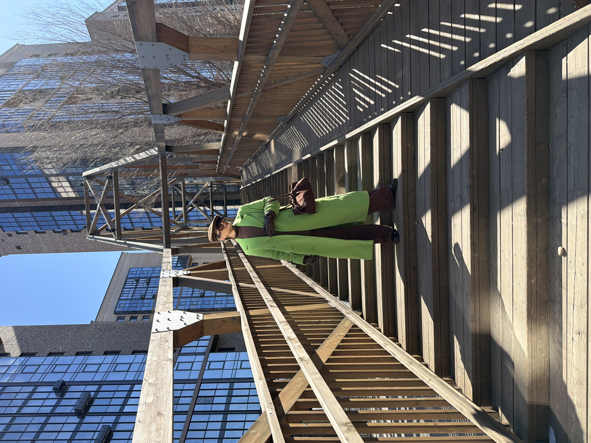 A photograph of a person wearing a bright green coat, brown pants, boots, gloves, and hat with a brown purse, appears to be standing on a wooden pedestrian overpass. The overpass features wooden slats for the walkway and is supported by large wooden beams, some of which are connected with metal plates. The sun casts strong shadows from the structure onto the walkway and the wooden siding of the overpass. In the background are buildings with glass windows, reflecting the clear blue sky. The picture is taken at an angle, with the buildings and sky visible in the upper left corner and the overpass receding into the distance on the right. The location is the pedestrian bridge at Tatyshev Island in Krasnoyarsk, Russia.