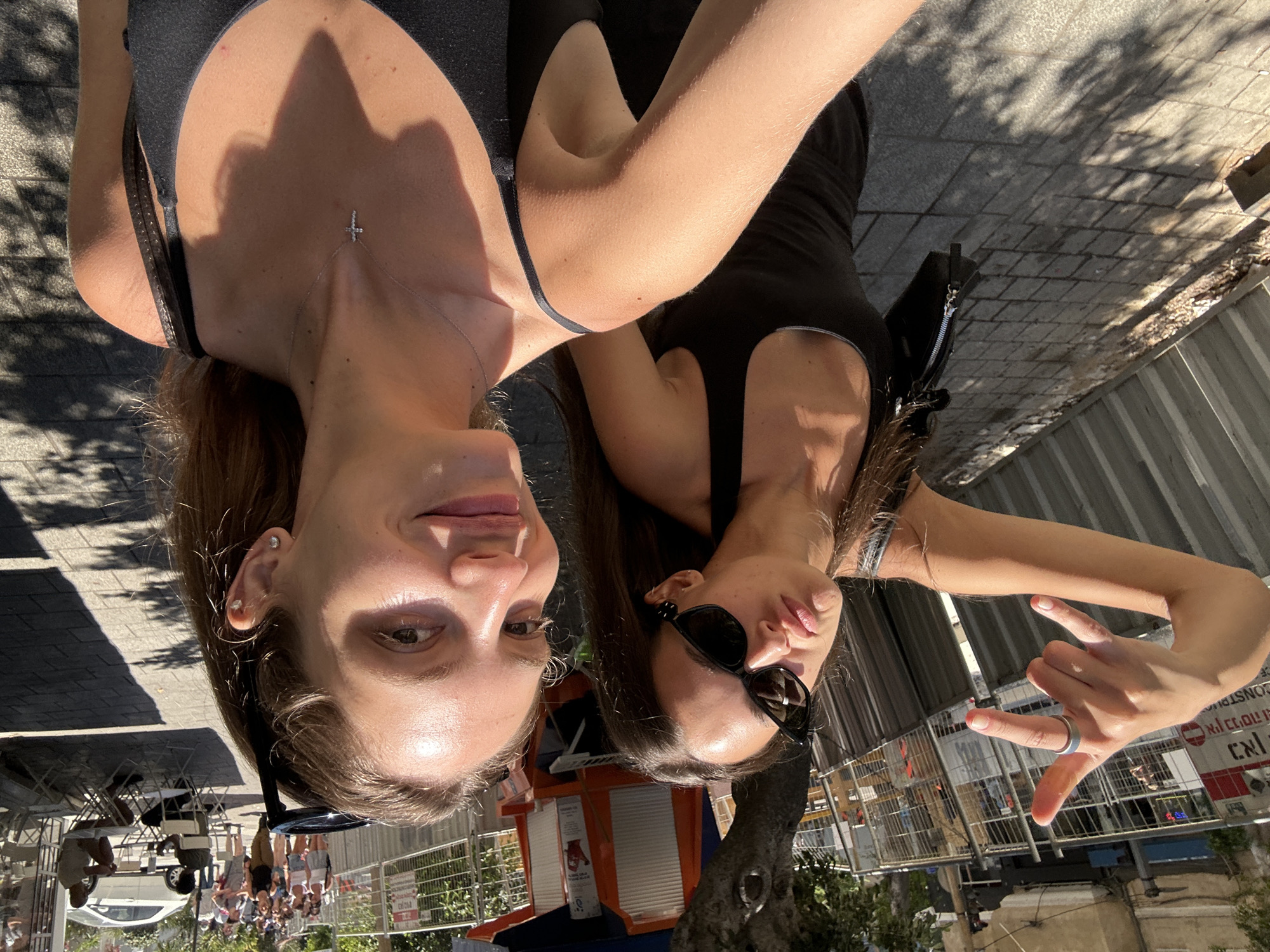 The image is a bright, sunlit selfie featuring two young women on an urban street, identified as Tel Aviv, Israel, due to visible Hebrew signage.
In the foreground, on the right, a young woman with light brown hair, pulled back by sunglasses perched on her head, smiles directly at the camera. She has visible freckles on her nose and cheeks and wears a dark gray or black spaghetti-strap top, a small silver cross necklace with clear stones, and a small silver stud earring in her right earlobe. Her right arm is partially visible in the lower right corner, likely holding the camera.
Slightly behind and to the left, a second young woman with long dark brown hair makes a playful, pouty face while wearing large black cat-eye sunglasses. She is wearing a dark, possibly black, tank top or dress. Her right hand is raised, making a "rock on" or "devil horns" gesture, with a silver band-style ring visible on her middle finger. A black shoulder bag with a silver zipper is slung over her left shoulder.
The background reveals a bustling street scene. To the immediate left, behind the second woman, is a construction or utility area enclosed by corrugated metal fencing and temporary gray metal barriers, featuring a sign with Hebrew text, including the word for "construction" (בנייה). Further back, trees provide shade, casting dappled shadows on the paved sidewalk, which is made of rectangular pavers.
To the right of the background, an active street is visible. Pedestrians are walking in the distance, and an outdoor café area with white tables and chairs is set up along the sidewalk, with two men seated at one table. A white sedan is visible on the street. A small, reddish-brown kiosk with a blue awning is situated in the mid-ground, beneath a tree. The overall scene suggests a warm, sunny day in a vibrant city environment.