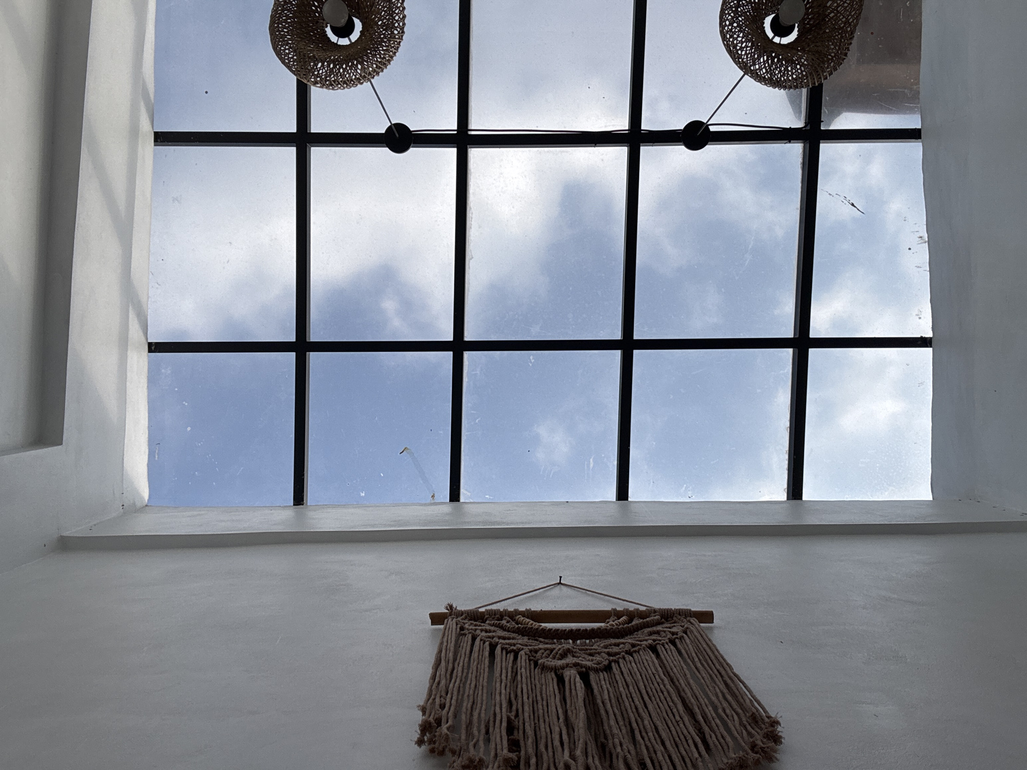 An interior view looking upwards shows a white wall on the left with a macrame wall hanging. To the right, a large, segmented skylight reveals a cloudy blue sky. Two woven pendant lights hang from the ceiling, one visible in the upper right quadrant and another in the lower right. The scene appears to be in a modern building, possibly a cafe or a home, located in the city of Kuta Selatan, Indonesia.