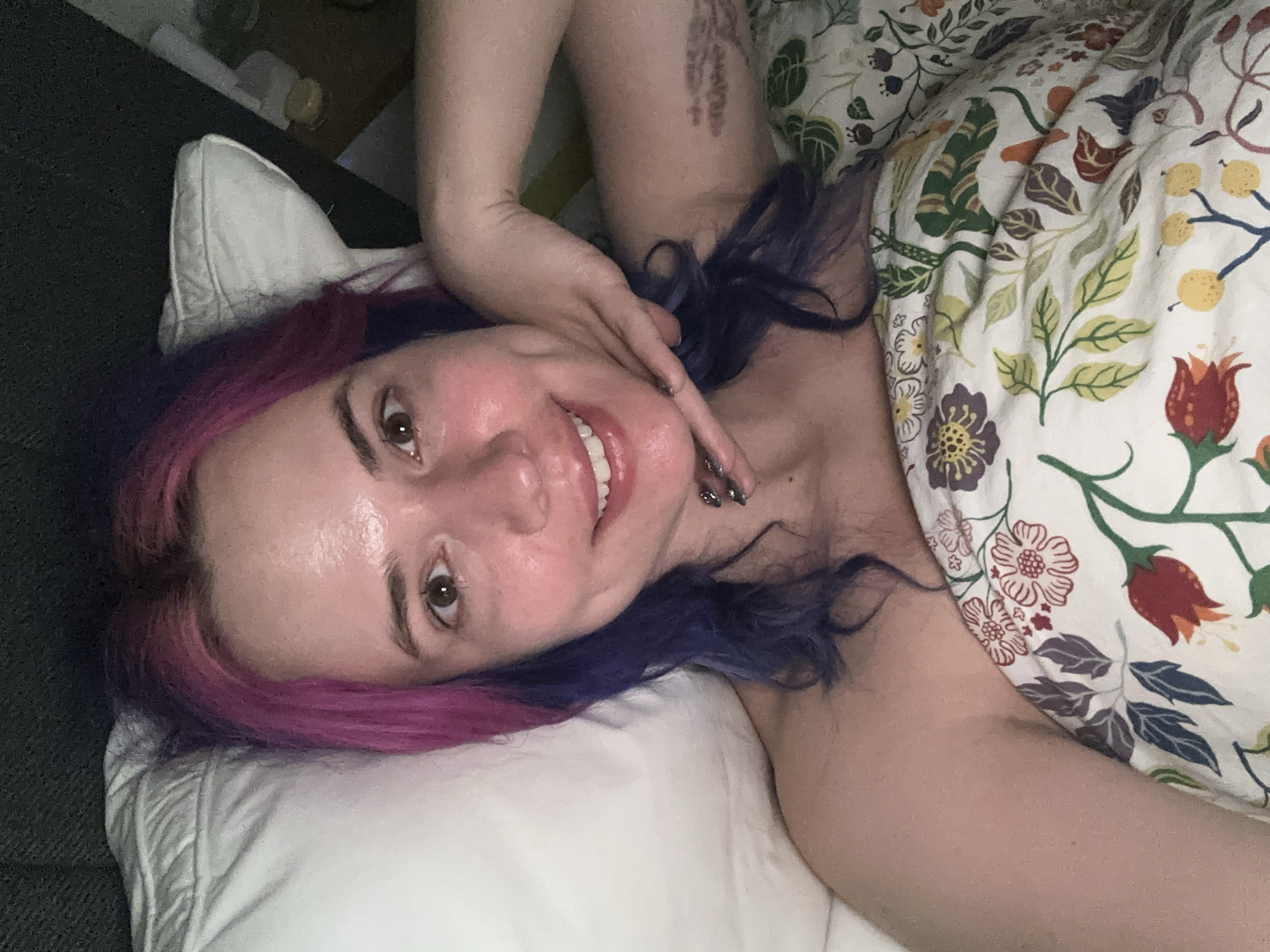 An indoor, close-up image captures a smiling person, likely female, lying down and looking directly at the camera, suggesting a selfie or posed photograph. The setting is identified as the City of Zagreb, Croatia. The person's head rests on a white pillow, and her hair is strikingly two-toned, featuring vibrant pink on one side and dark blue or purple on the other. She has a contented or happy expression, with her mouth slightly open in a smile, revealing white teeth. Her right hand is positioned near her chin and neck, showcasing fingers with dark nail polish, and a tattoo is visible on her right upper arm.
She is partially covered by a colorful floral blanket or duvet, which displays an intricate pattern of leaves and various flowers in shades of green, red, yellow, purple, and orange. To the left side of the frame, dark grey or black furniture, possibly a couch or a headboard, is visible. The lighting appears soft and even, characteristic of an indoor environment, but does not provide specific cues about the time of day or outdoor weather. There is no visible text in the image.