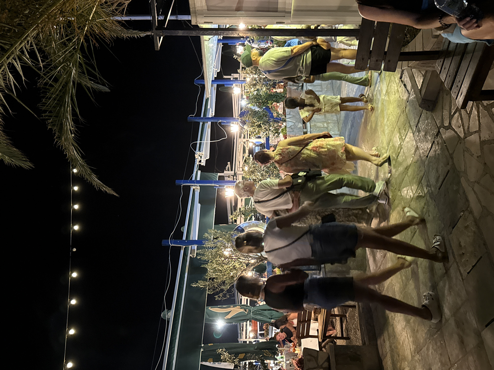The image depicts an outdoor dining area at night, likely in Budva, Montenegro. String lights are visible overhead, illuminating a paved walkway and seating areas. Several people are present, some walking and others seated at tables. A group of individuals, including two women in denim shorts and two men in casual attire, are walking away from the camera. One man is wearing a green cap and shorts, while another is in a white shirt and light-colored pants. A young girl in a white outfit is also visible. The background features outdoor seating with green umbrellas, plants, and building structures, suggesting a restaurant or cafe. The overall atmosphere is relaxed and evening-oriented.