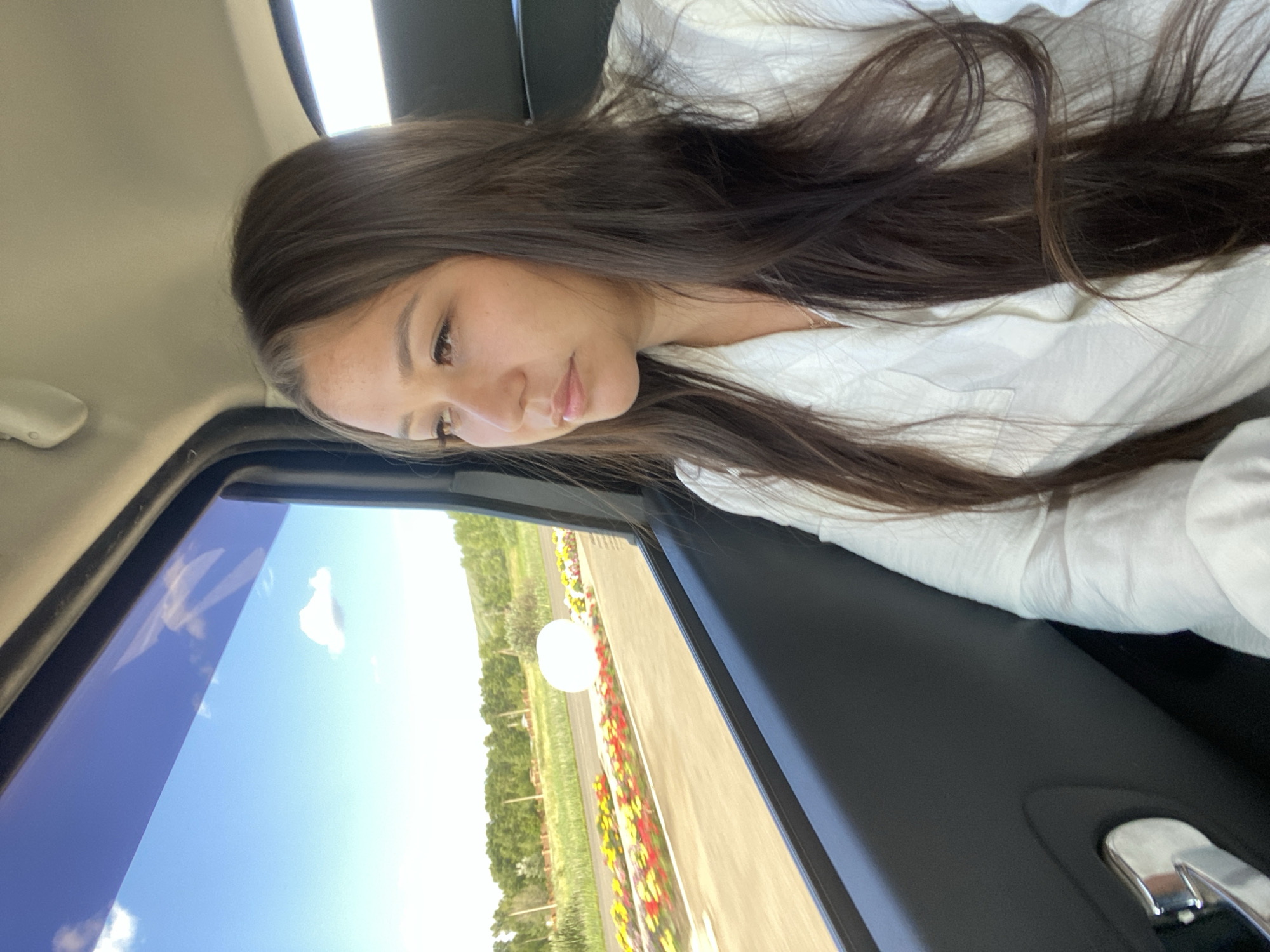 A young woman is seated in the passenger seat of a car, looking out the window. The woman has long, dark brown hair and is wearing a white button-up shirt. The car's interior is visible, with the dark gray door panel and a silver door handle in the foreground. Outside the window, a bright blue sky with scattered white clouds is visible, along with a lush green landscape of trees and a field of colorful flowers lining a road. The setting appears to be a scenic area, possibly in or near Isyangulovo, Russia, given the rural scenery. The image captures a moment of quiet observation as the woman travels.