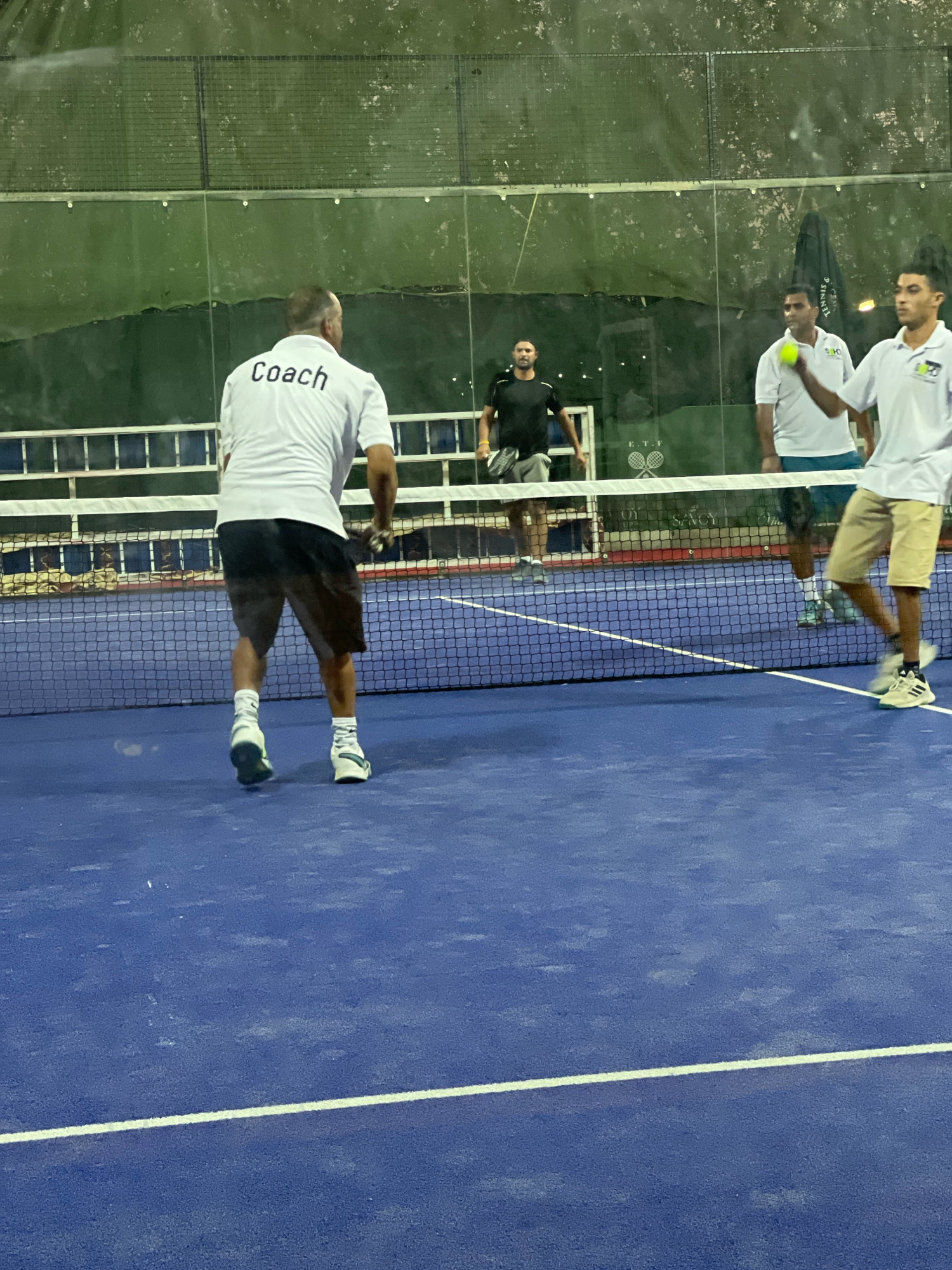 Four men are playing padel tennis on a blue court in Sharm El Sheikh, Egypt. In the foreground, a man wearing a white shirt with "Coach" printed on the back and dark shorts is facing away from the camera, positioned near the net. To his right, on the other side of the net, three men are visible. One man in a white shirt and blue shorts is holding a yellow padel ball. Another man, also in a white shirt, is to his right, and a man in a black shirt and grey shorts is on the far left, holding a padel racket. The court is enclosed by glass walls and netting, with trees and structures visible in the background. The lighting suggests it is either dusk or an indoor court with artificial lighting.