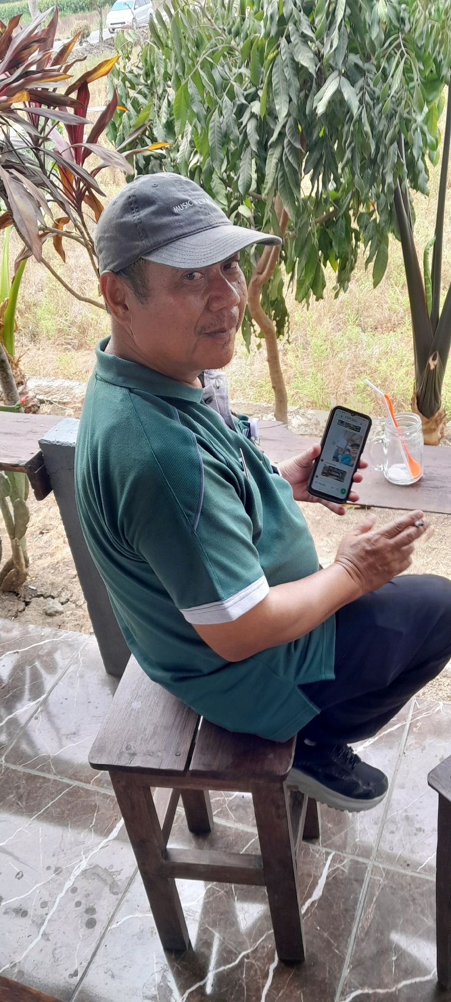 A man wearing a green polo shirt and a grey baseball cap sits on a wooden stool on a tiled patio. He holds a smartphone in his right hand and a cigarette in his left. A glass with a straw and what appears to be ice and a powder is on a nearby wooden table, along with the corner of another stool. Lush green foliage and trees surround the man, suggesting an outdoor setting. The man appears to be looking towards the camera. The environment is consistent with a relaxed, outdoor area, possibly a cafe or home patio in Sidohasri, Indonesia, given the presence of tropical vegetation.
