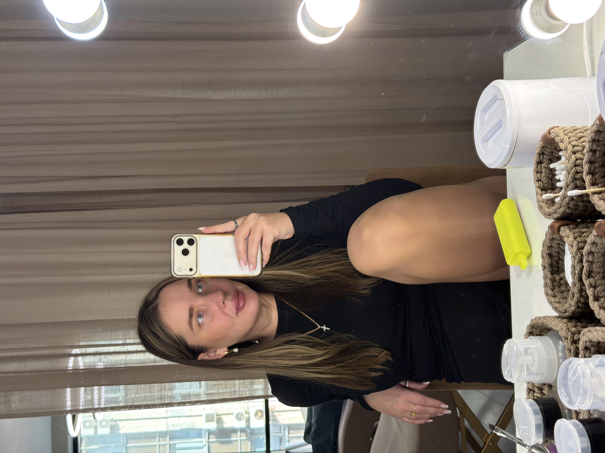 A person with fair skin and light brown hair, wearing a black long-sleeved shirt and a silver cross necklace, takes a selfie in front of a mirror. The reflection shows the person holding a white and gold smartphone with the camera facing the viewer. The mirror has bright lights along the top. In the background, there are beige curtains and a view of buildings through a window, suggestive of an urban environment. To the right of the person in the mirror, there are a series of white jars, brown knitted containers holding cotton swabs, and a yellow container on a white surface. While it's not possible to definitively pinpoint the location as Dolgoprudny, Russia, the architectural style and environmental features visible through the window are consistent with urban areas in Russia.