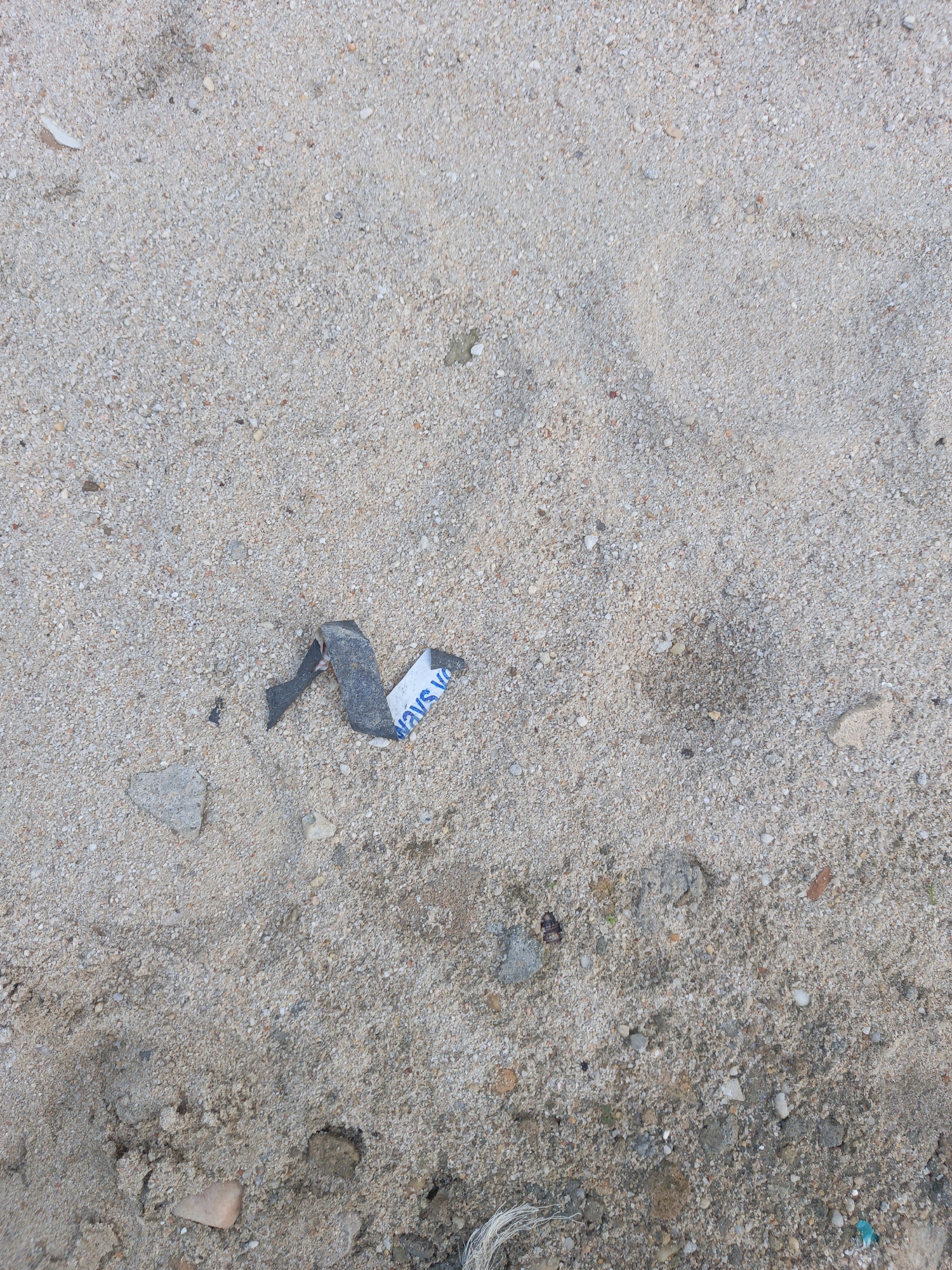 The image shows a patch of coarse sand, likely on a beach or in a sandy area in the city of Unknown, Unknown. Embedded in the sand are small pebbles, bits of shell, and debris. A torn piece of tape or sticker with blue writing on a white background is visible toward the middle of the image. A dark, possibly metallic piece is attached to the tape/sticker. At the bottom of the image, a bit of white string or thread is seen.