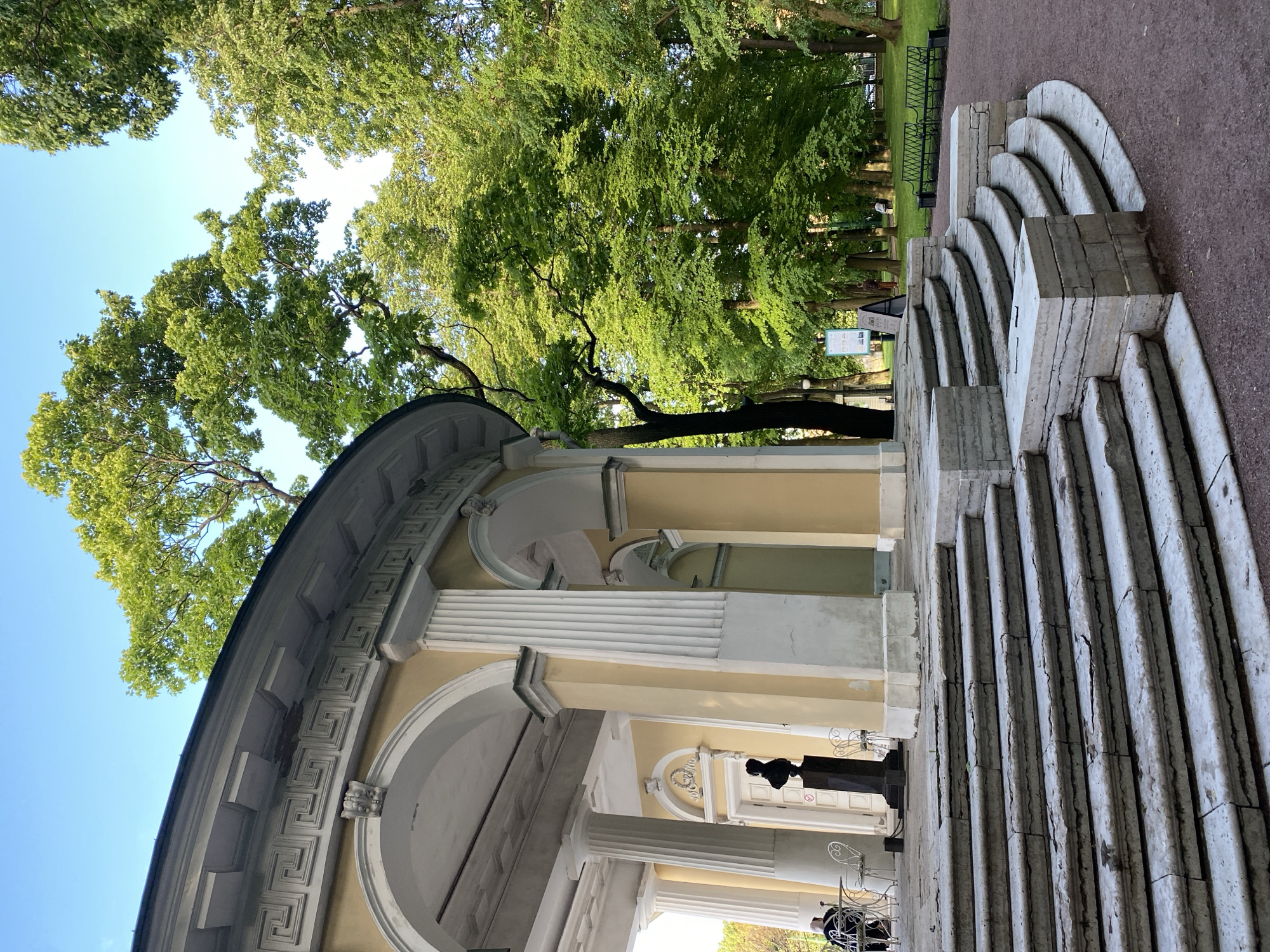 The image shows the Rotunda in the Tauride Garden, Saint Petersburg, Russia. The Rotunda, a semicircular pavilion with classical columns and a decorative frieze, is painted in a pale yellow hue. It is situated at the top of a grand, semicircular stone staircase, which is in a state of disrepair with visible cracks and chips on the steps.
In front of the Rotunda, on the landing of the staircase, stands a dark bust on a pedestal. To the left, a person is seated at a small white table with a white chair. Beyond the Rotunda, the Tauride Garden unfolds, characterized by lush green trees, a clear blue sky, and pathways. Several park benches are visible in the background, and a few indistinct figures can be seen in the distance, enjoying the park. The overall scene depicts a historical architectural structure within a serene park setting on a bright day.