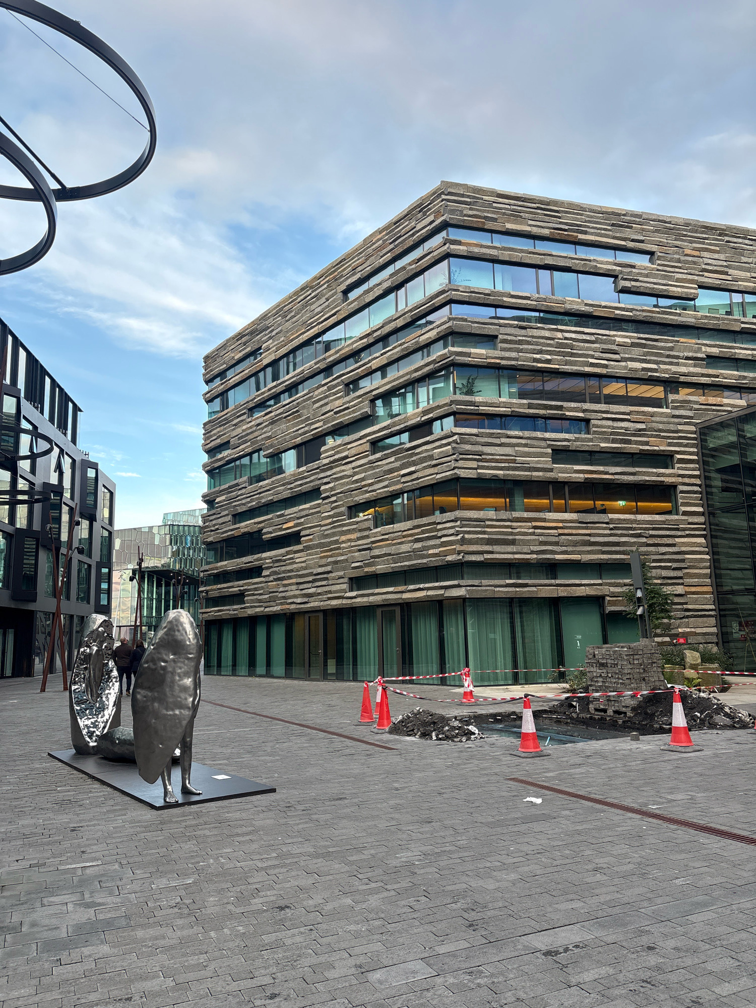 In Reykjavik, Iceland, an outdoor public space features modern architecture and public art. A large, multi-story building with a facade of horizontal stone slabs and extensive glass windows dominates the right side of the frame. Adjacent to it on the left is another modern building with dark, geometric window frames. In the foreground, two abstract, metallic sculptures are displayed on a dark rectangular base. Two individuals stand near the sculptures, facing away from the camera. The ground is paved with grey rectangular stones. To the right, construction is underway, with orange and white traffic cones, piles of dirt, and barriers indicating a disturbed area. The sky above is a mix of blue with scattered clouds.