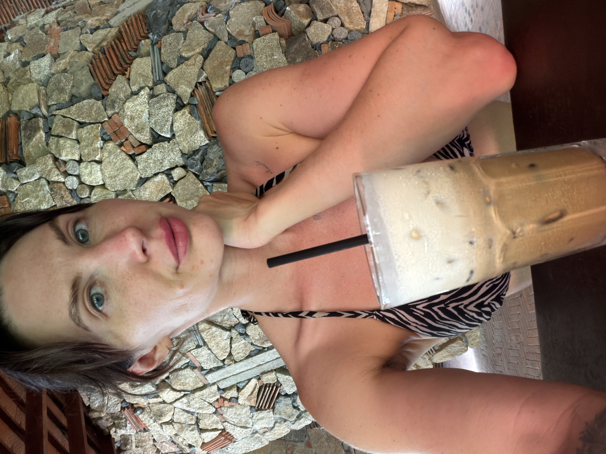 A young Caucasian woman with brown hair and blue eyes is captured in a close-up selfie, holding a glass of iced coffee. She is wearing a black and white patterned bikini top. Her left hand rests on her chin, and her right arm is bent and resting in front of her chest. The background features a wall constructed of rough, uneven stones and terracotta tiles. The scene appears to be outdoors, with wooden beams visible in the upper left corner, suggesting a covered seating area or pavilion. The lighting suggests it is daytime. The setting is consistent with an outdoor cafe or restaurant in Tay Nha Trang Ward, Vietnam, possibly near a beach or resort, given the attire and the natural, rustic background.