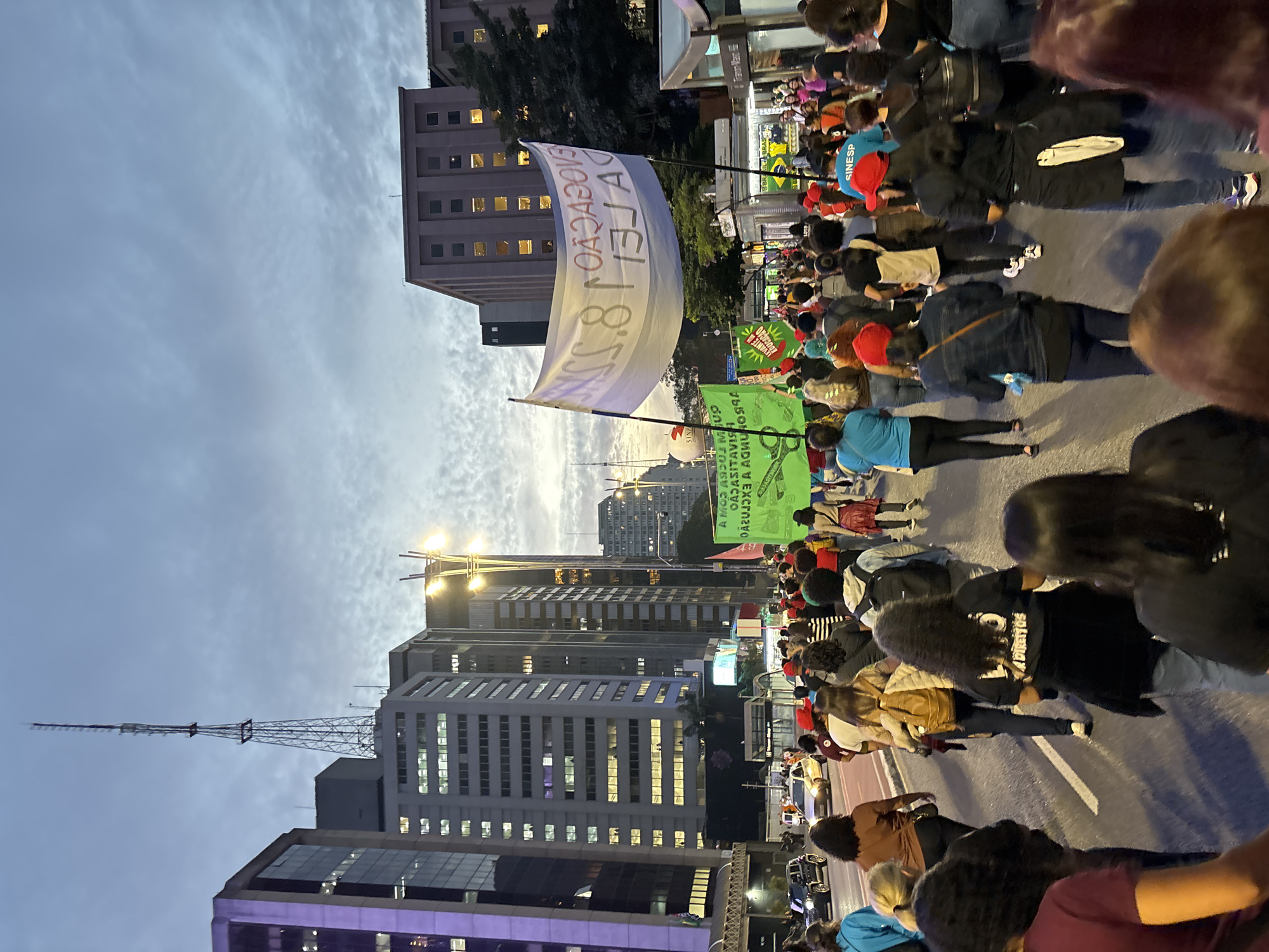 Here is a description of the image:
A large crowd of people are marching down Avenida Paulista in São Paulo, Brazil. The march appears to be a protest or demonstration, as many participants are holding signs and banners. One large white banner at the front of the crowd reads "LEVANTE DA EDUCAÇÃO." There are other signs as well, some of them green and one saying "LOTO". The people are dressed in a variety of casual clothing. Buildings line the street, including some modern high-rise structures with many windows lit up. A tall radio or television tower is visible. The sky is partly cloudy, and it appears to be either dusk or dawn.
