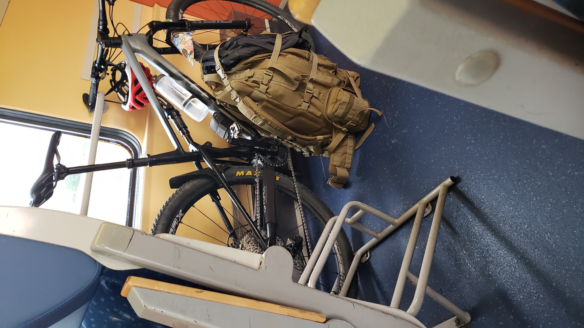 The image shows a bicycle parked inside a train car. The bicycle is a full-suspension mountain bike with a gray frame, black handlebars, and black wheels. A tan backpack is leaning against the bicycle, and a red and white helmet is attached to the handlebars. The train car appears to be empty, with blue seats visible in the foreground and a yellow wall in the background. The floor of the train car is a dark blue speckled material. The lighting suggests it is daytime, and the window in the background shows a blurred outdoor scene. The location is identified as Sučany, Slovakia.