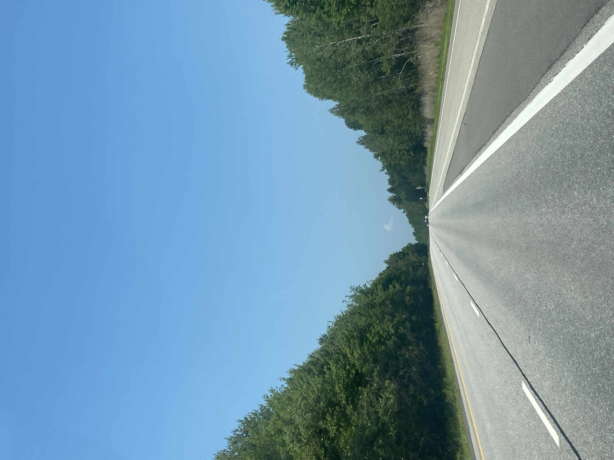 The image shows a highway stretching into the distance, flanked by dense green trees on both sides. The sky is a clear, bright blue, suggesting a sunny day. The road appears to be asphalt with white lane markings. The perspective is from a vehicle moving forward, as indicated by the vanishing point of the road. The overall scene evokes a sense of travel and open space, with the vibrant green of the trees contrasting with the gray of the road and the expanse of the blue sky. There are no people or specific objects visible that would indicate a particular event. The time of day appears to be daytime, likely late morning or afternoon due to the bright sunlight. The location is East Hawkesbury, Canada.