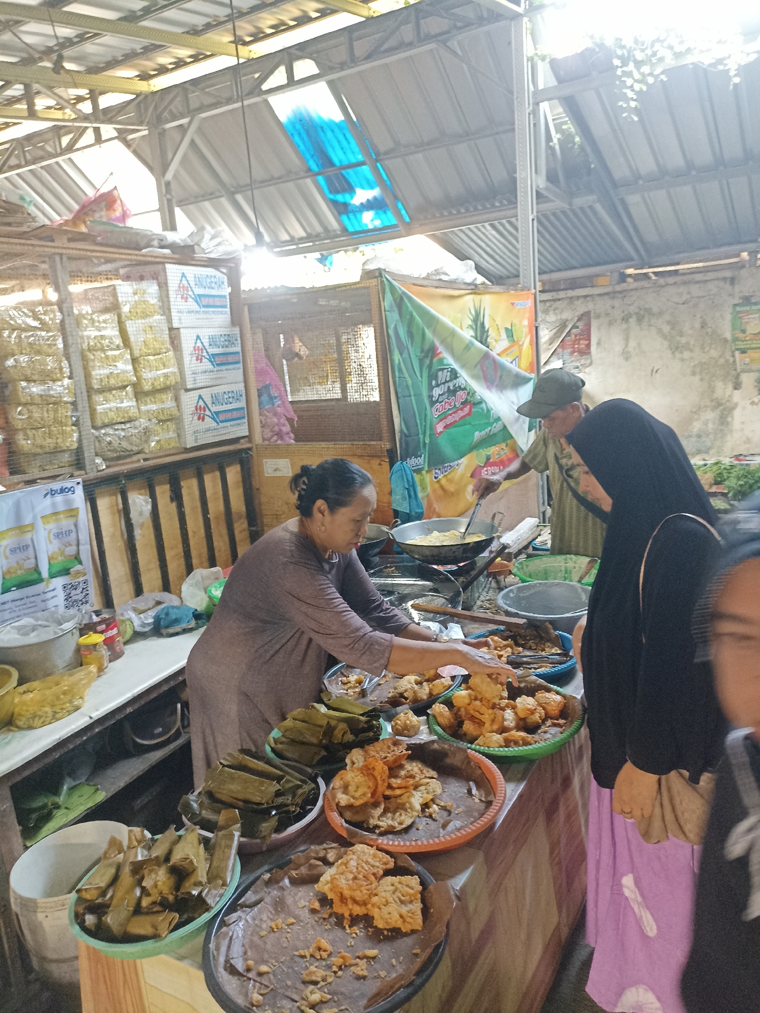The image shows an outdoor food stall, likely in Kelurahan Kepatihan, Indonesia. A woman in a brown shirt is preparing food, possibly frying it in a large pan over a stove. She is surrounded by various food items displayed in bowls and on platters, including fried snacks and wrapped parcels. Two women, one wearing a black hijab and the other a purple skirt, are observing or interacting with the food. In the background, there are shelves stocked with bagged goods, a banner advertising a food product, and a corrugated metal roof structure. The scene depicts a bustling market environment where food is being sold and prepared.