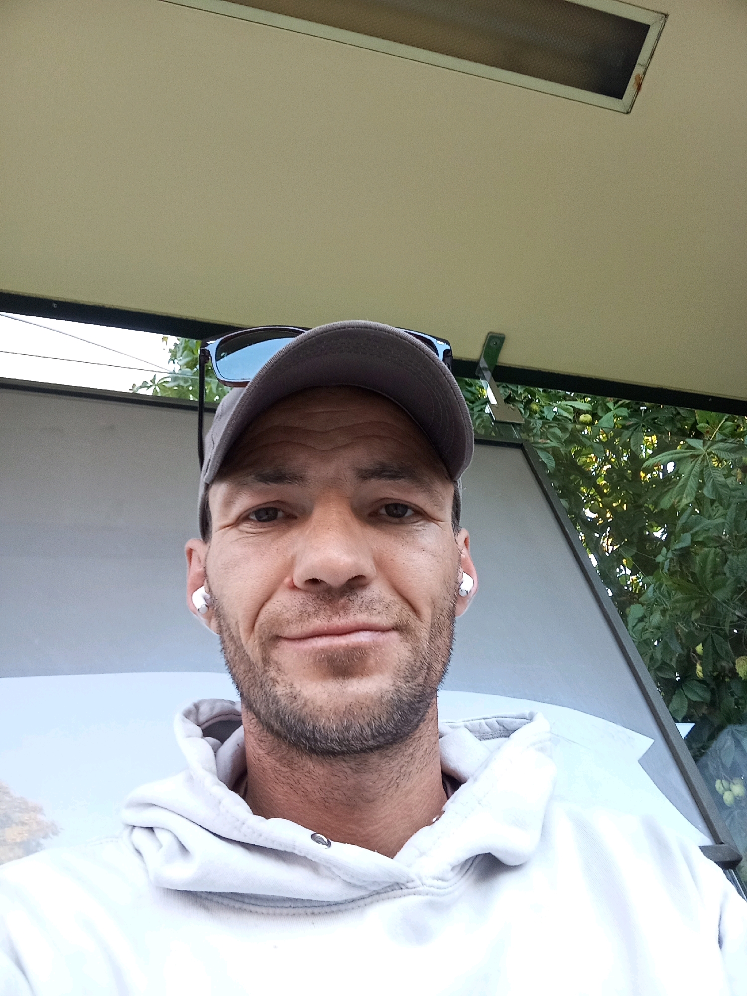 A man with short brown hair, a beard, and a light-colored hoodie is taking a selfie outdoors. He is wearing a grey baseball cap with sunglasses perched on top. He also has white earbuds in both ears. The background includes green foliage, suggesting a natural or park-like setting. The overall lighting suggests it is daytime. The location is not discernible from the image.
